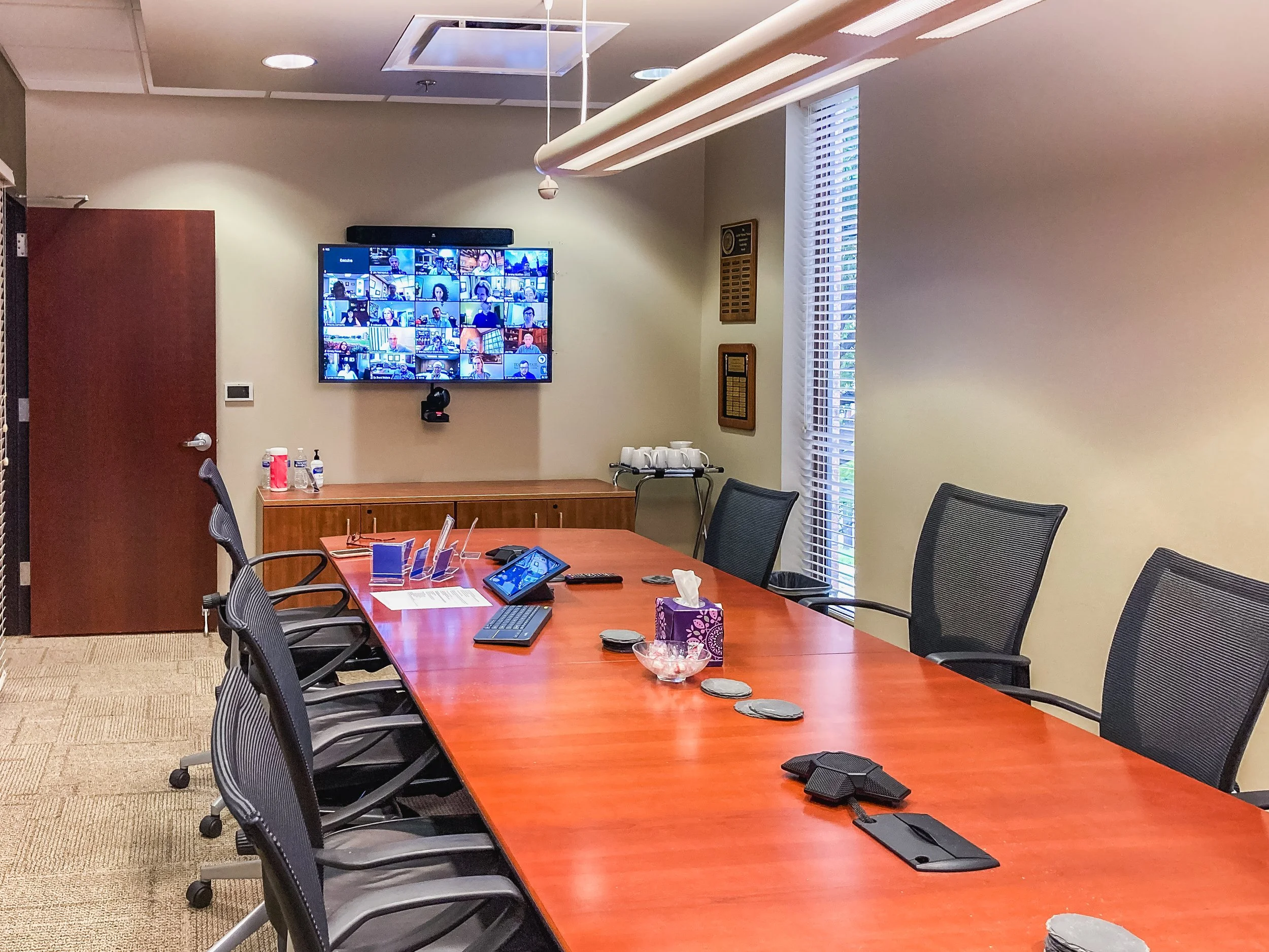 A conference room with a long wooden table surrounded by black mesh office chairs. The table has various items including a tablet, tissue box, and coasters. A wall-mounted TV screen displays a video call with multiple participants. The room has beige