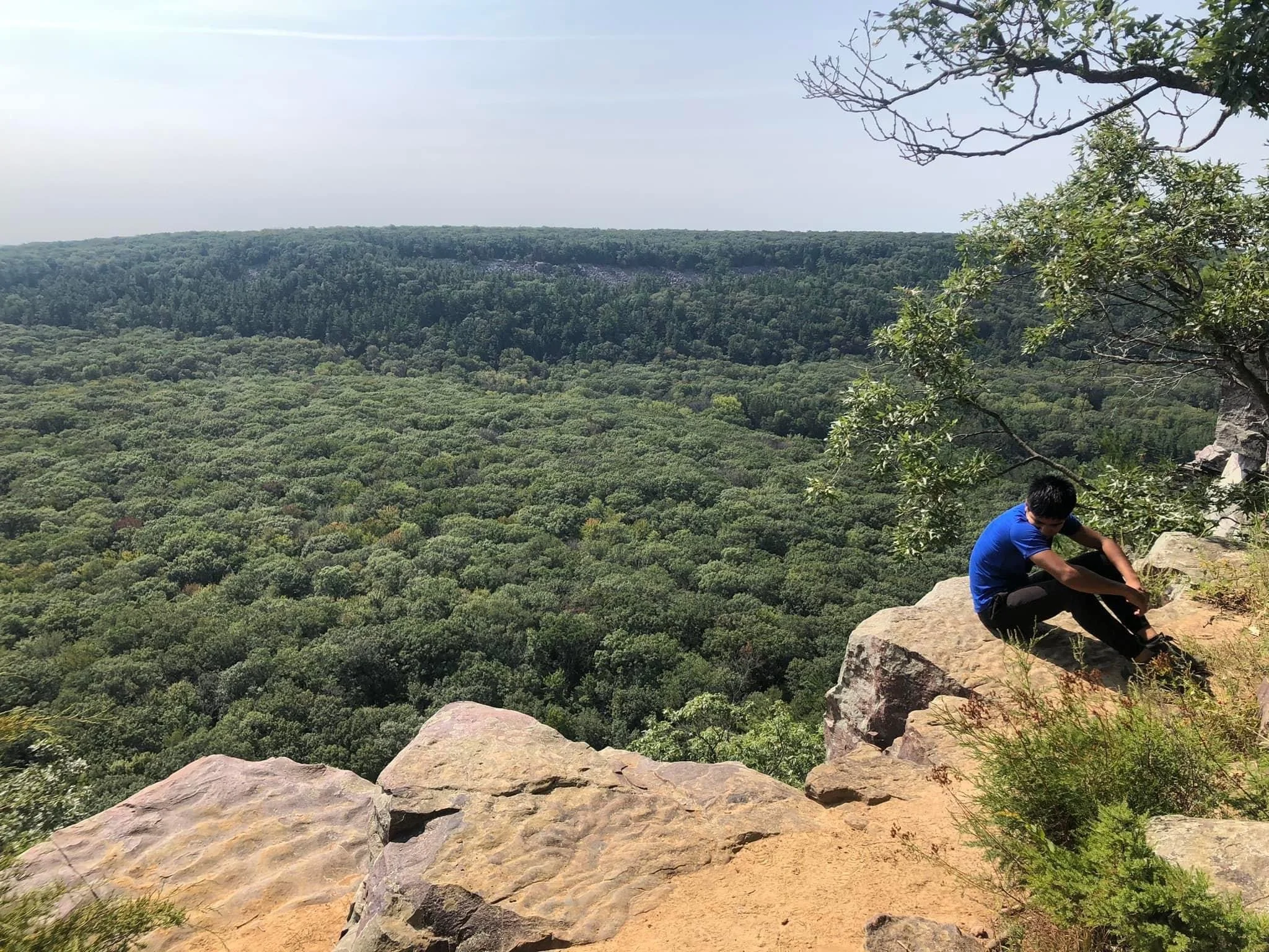 Person sitting on rocks at edge of cliff overlooking a green forested canyon and distant hills under cloudy sky.
