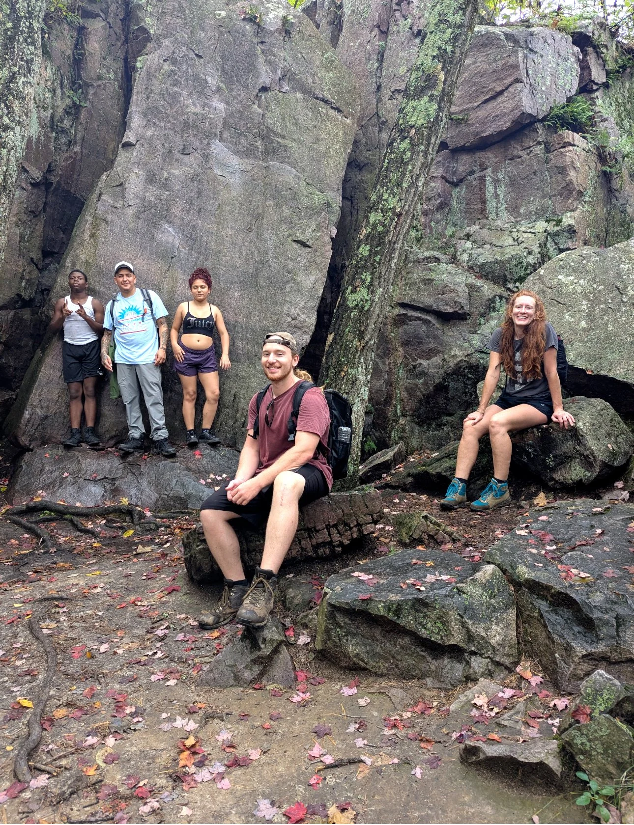 Five hikers in a forest with large rocks and trees, two sitting and three standing, smiling and posing for the camera.