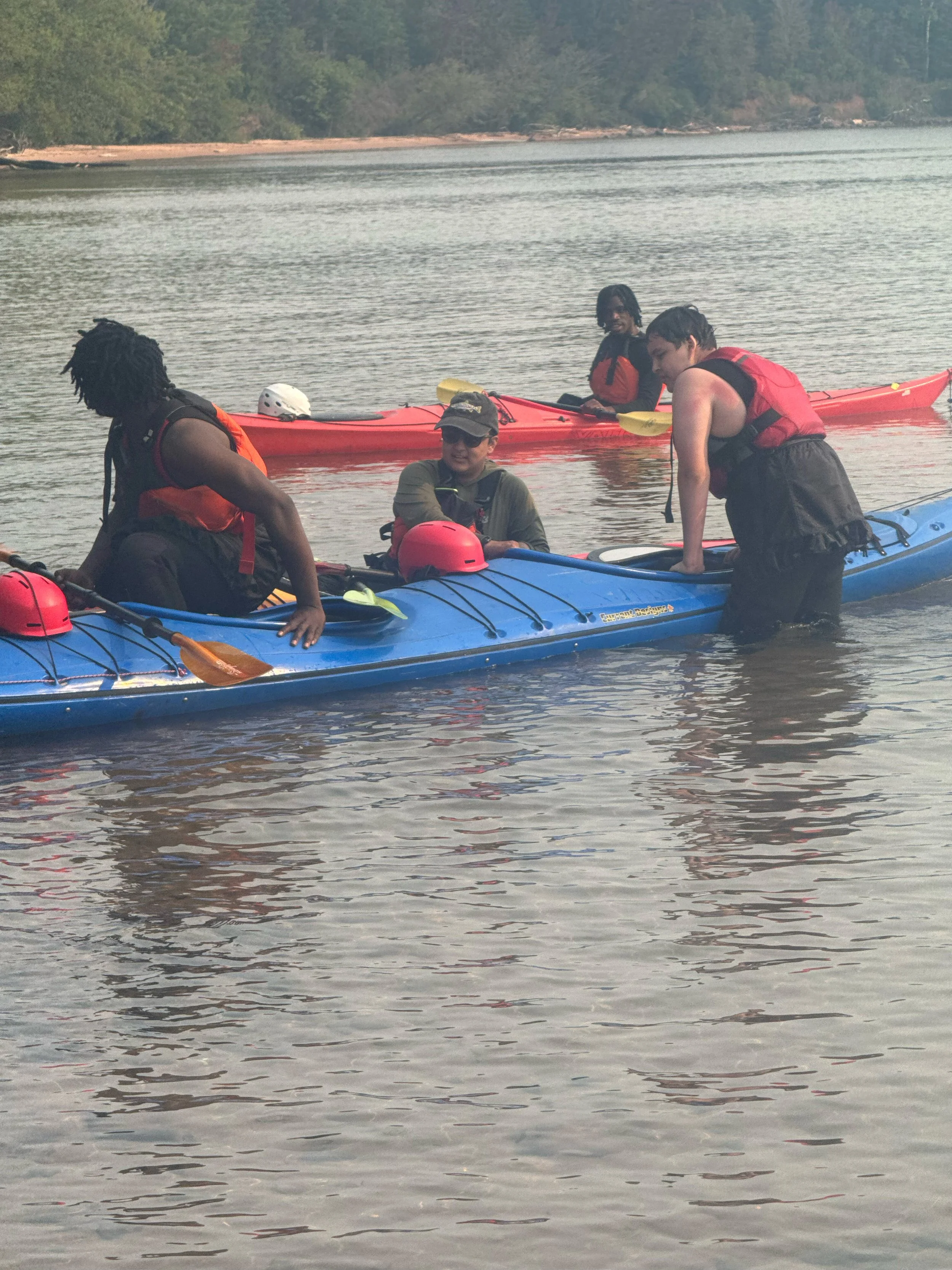 Four people in life jackets with kayaks on a calm lake, surrounded by trees.