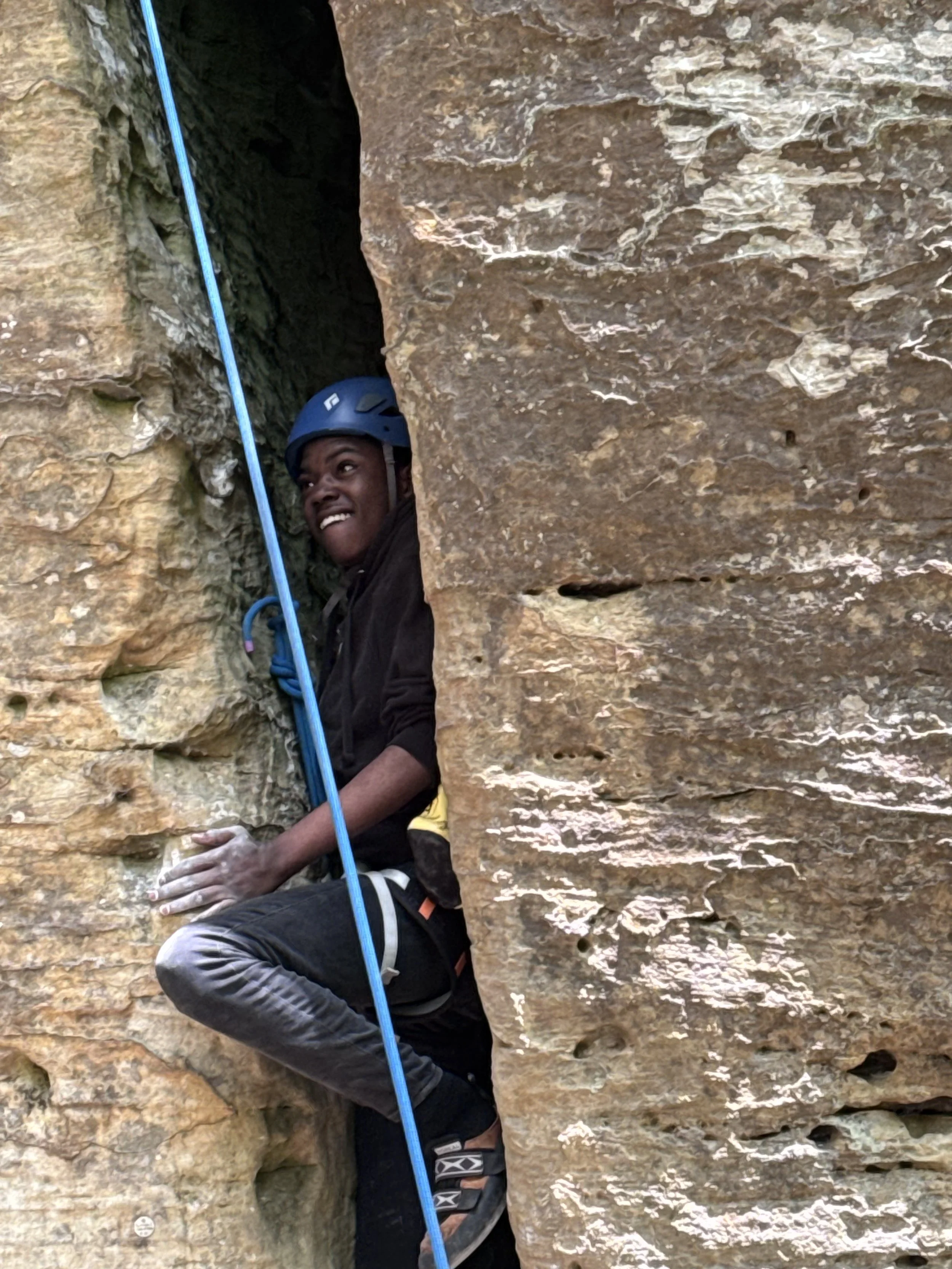 A person wearing a black helmet and black clothing rock climbing on an outdoor rock face with a blue climbing rope.