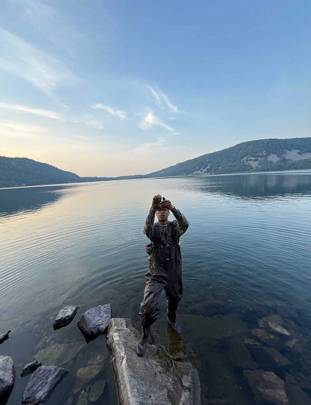 A man standing on rocks in a lake, with mountains and a cloudy sky in the background.