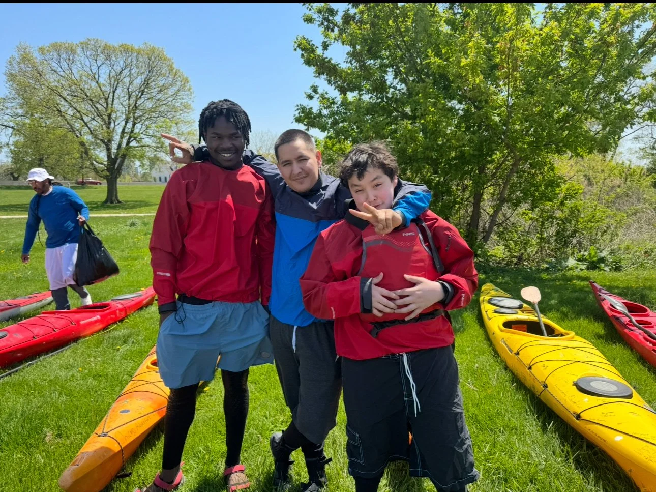 Three smiling kids posing with kayaks outdoors on grass on a sunny day with trees in the background, and a man walking away with a bag.
