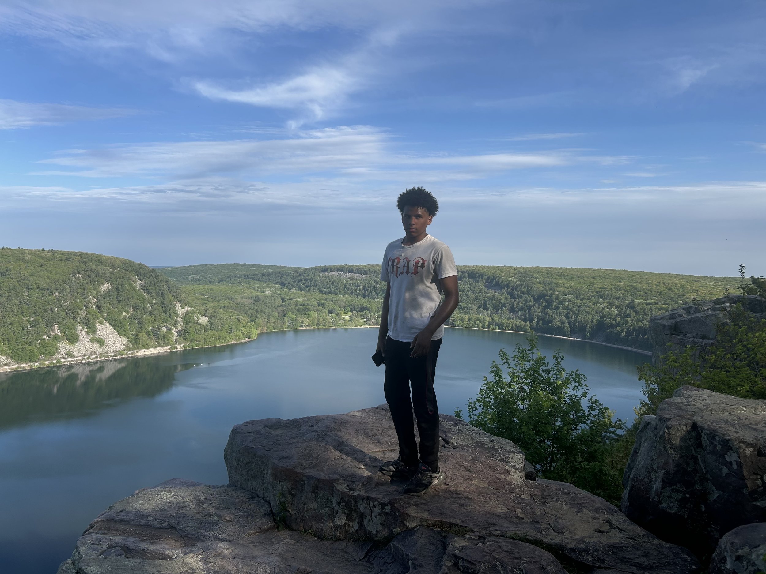 A young man standing on a large rock overlooking a river, with lush green hills and a partly cloudy blue sky in the background.
