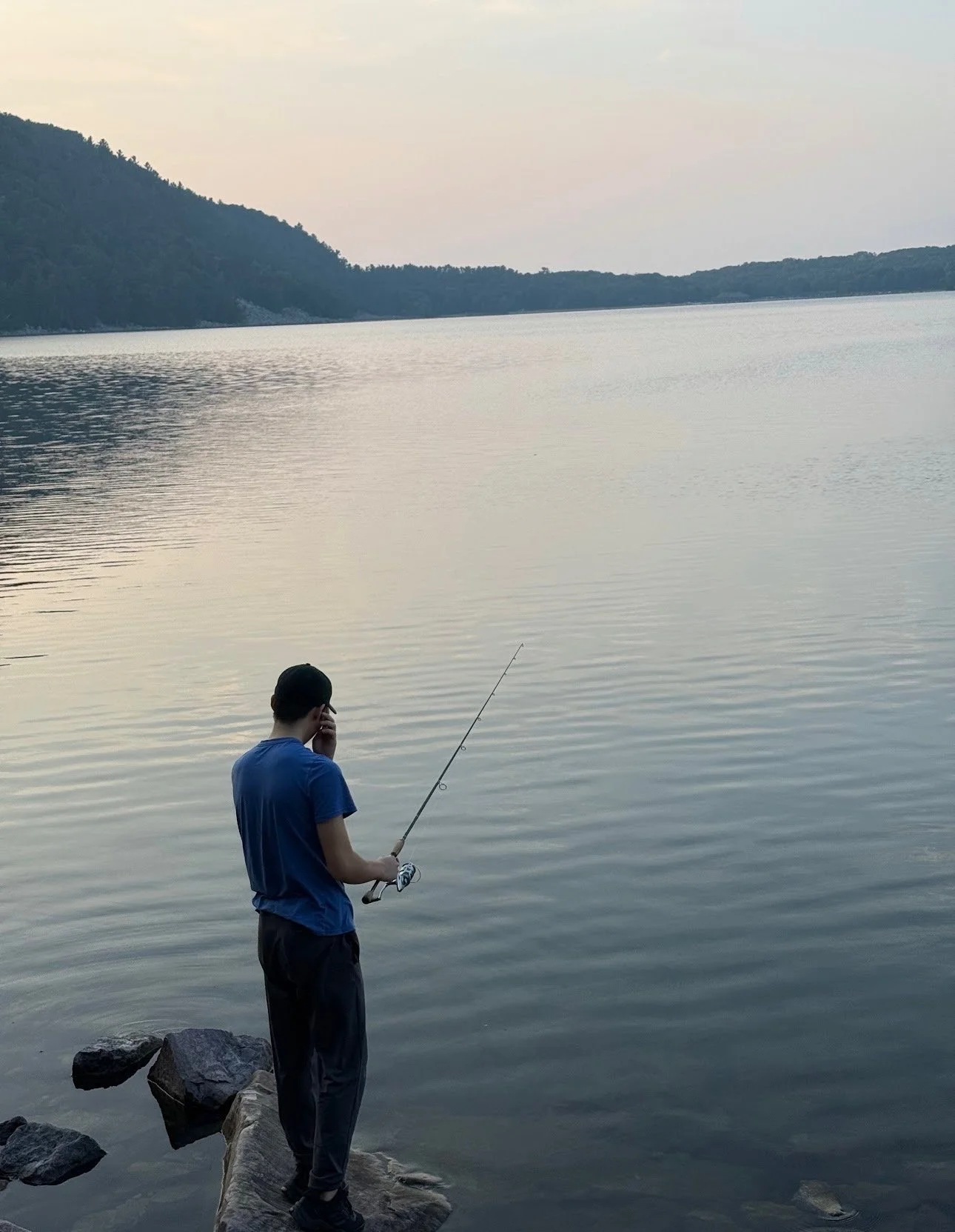 A person standing on rocks at the water's edge, fishing with a rod and reel, looking at the lake, with hills in the background during sunset.