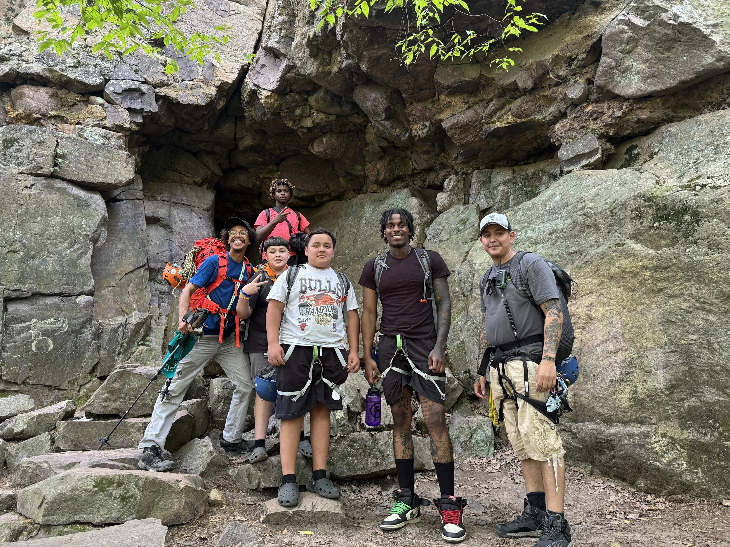 Group of seven hikers standing on rocky terrain in front of a large rock formation in a forested area. They are smiling, with some making peace signs, dressed in outdoor gear and carrying backpacks.