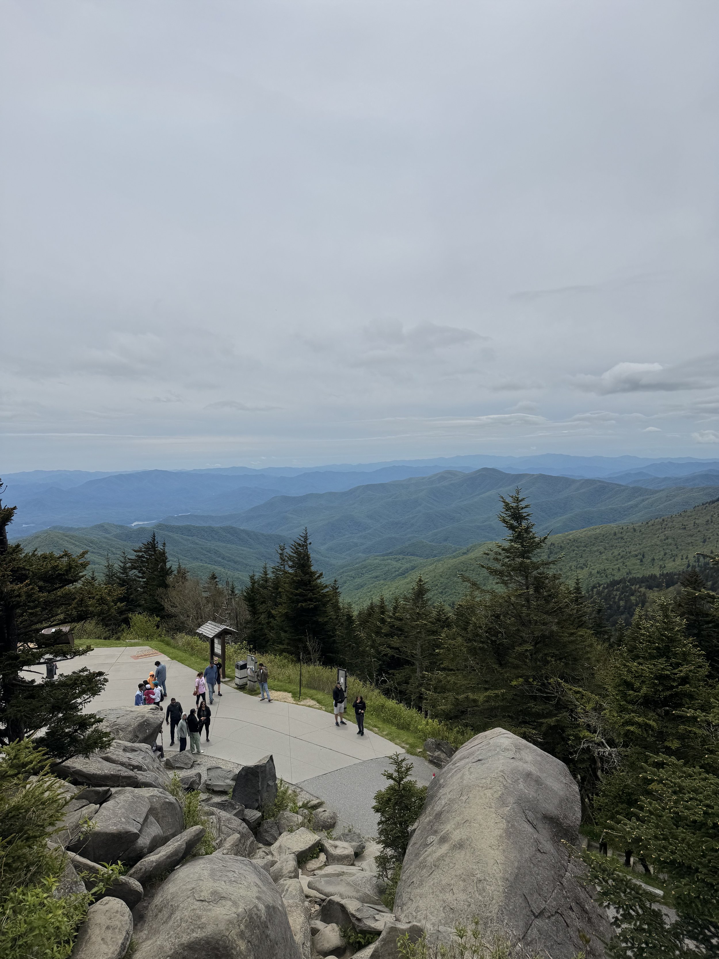 Scenic view of a mountainous landscape with rolling green hills and trees, and a group of people walking on a paved trail in a park.