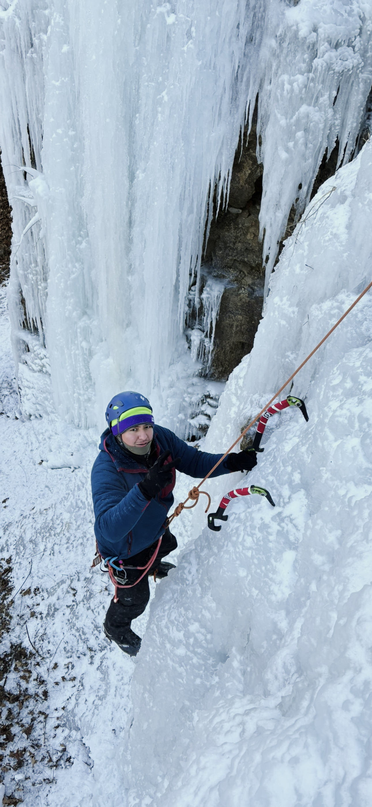 Climber in a blue jacket and helmet standing next to a frozen waterfall, holding ice axes and making a peace sign.