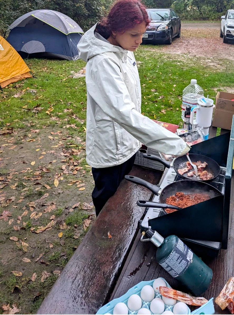 A person cooking outside on a camping trip. They are frying meat in a pan, surrounded by camping tents and parked cars.