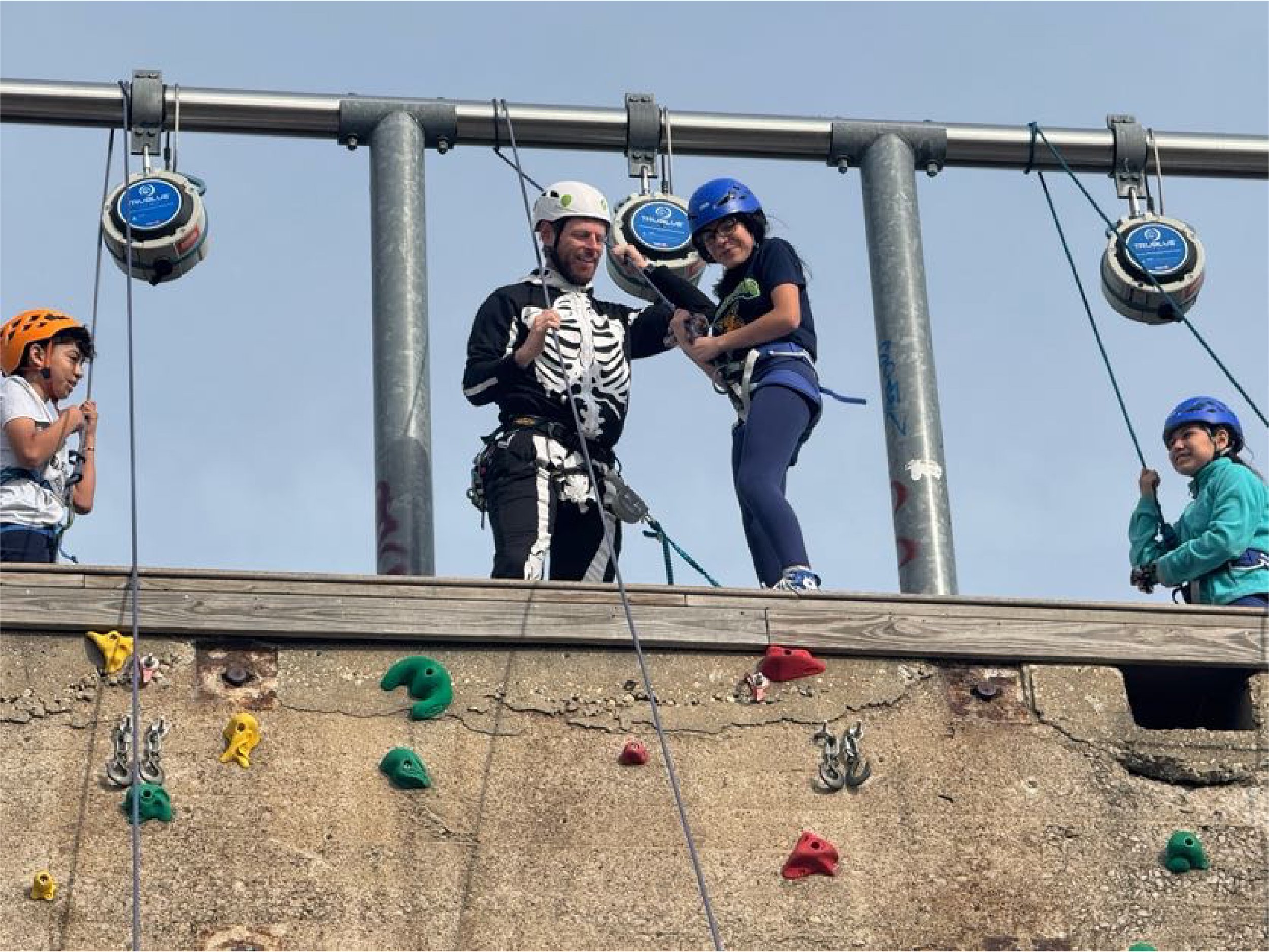 People wearing climbing helmets and harnesses on a high ropes course, with colorful handholds on a vertical wall below.
