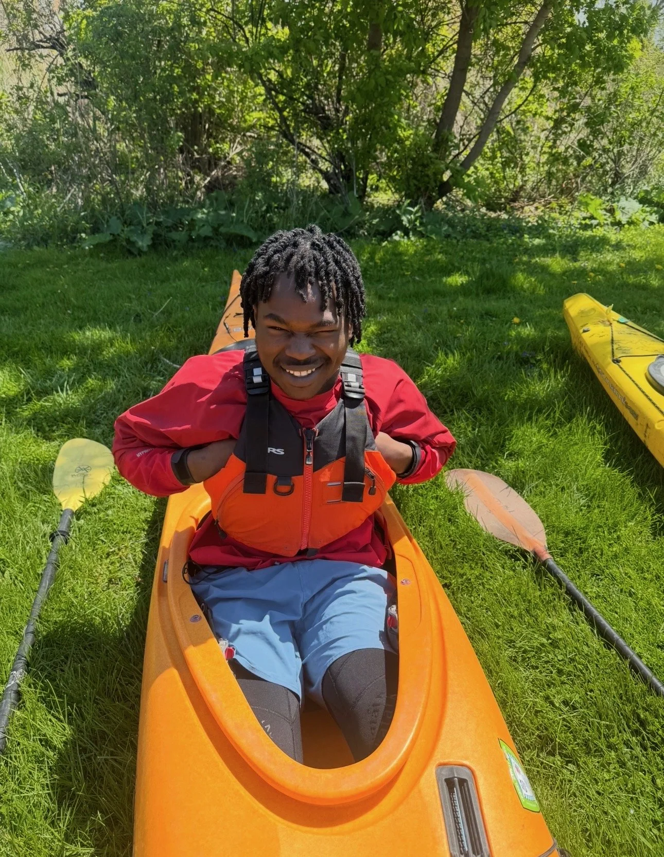 A man smiling while sitting in an orange kayak on green grass, surrounded by trees, with a yellow kayak and paddles nearby.