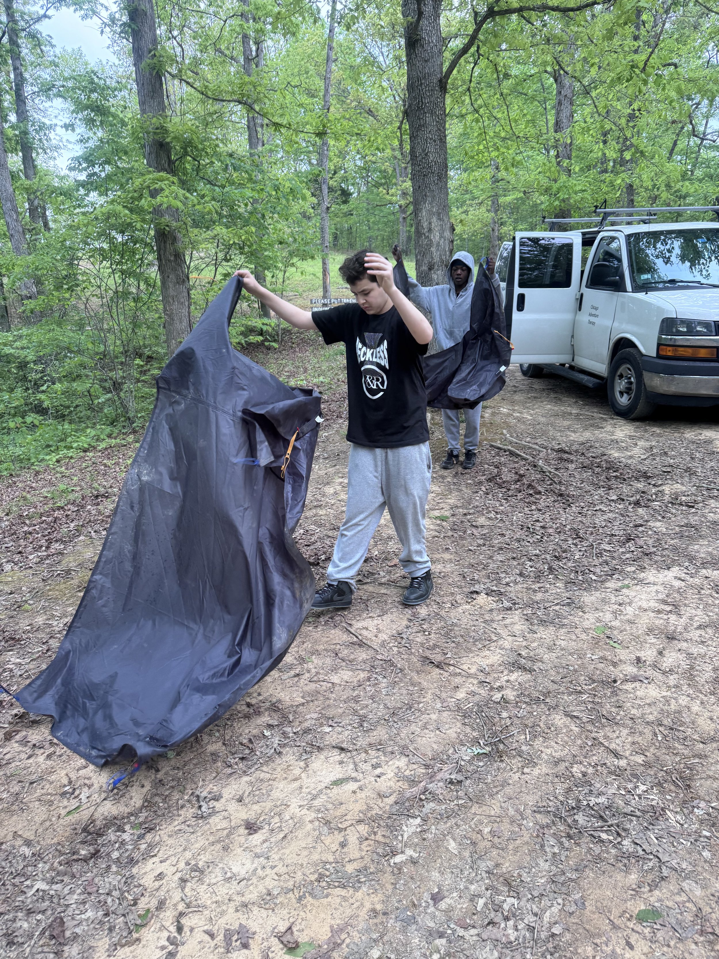 Two boys in a wooded area packing up or setting up a tent with a white service van nearby.