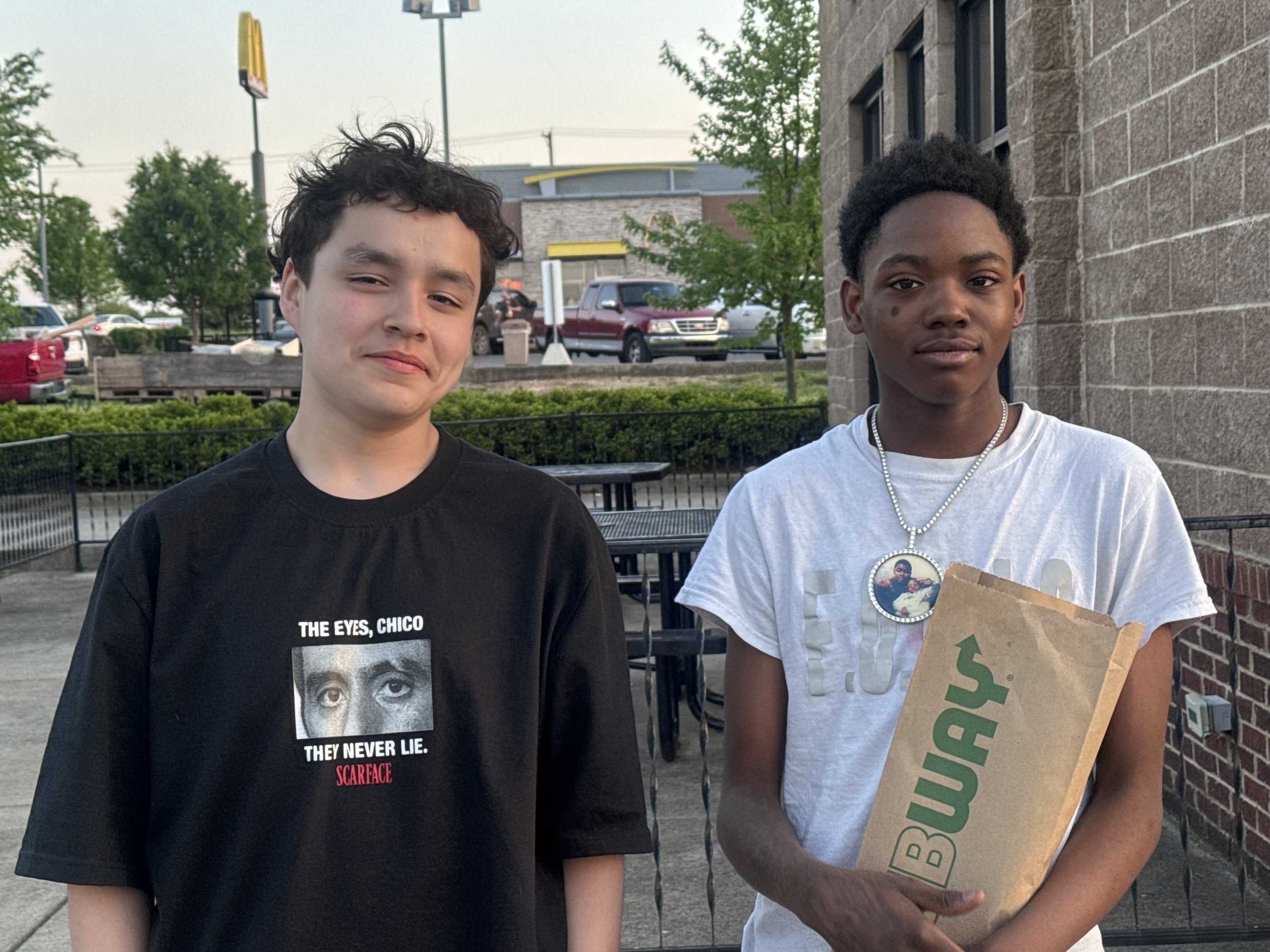 Two young men standing outside near a brick building and parking lot, one in a black t-shirt with a photo of a person's face and the words "The Eyes, Chico" and the other in a white t-shirt with a photo and a necklace, holding a pizza box from Subway.