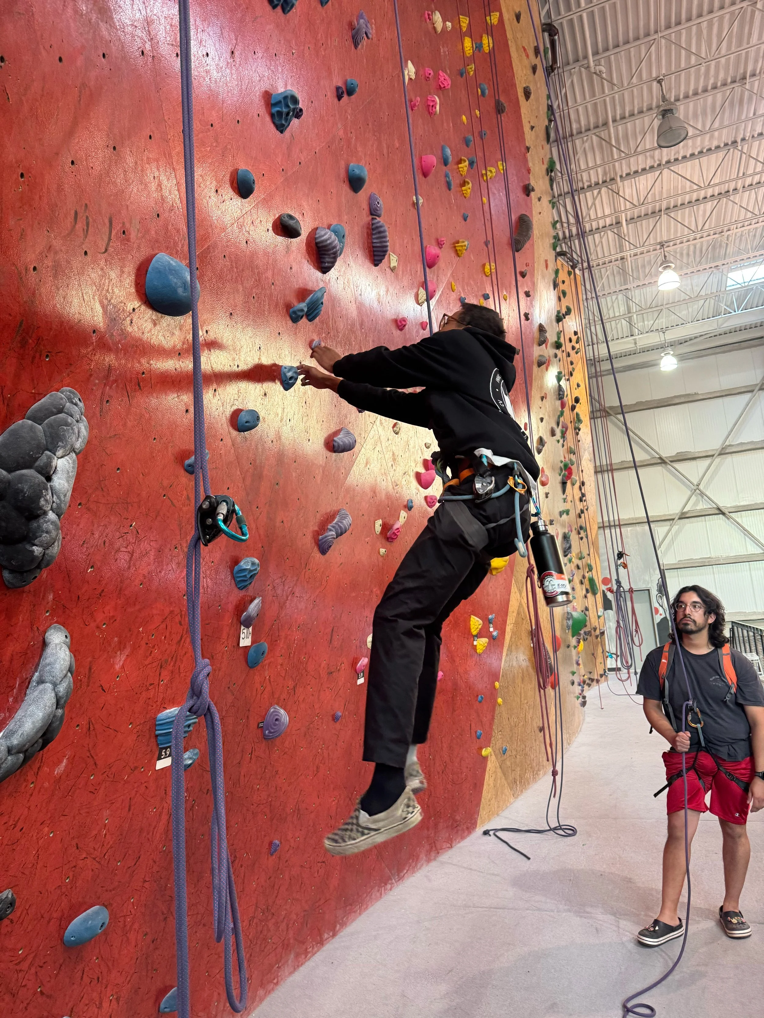 People bouldering at an indoor rock climbing gym with climbing holds on artificial red wall and someone tying in or preparing their climbing harness.