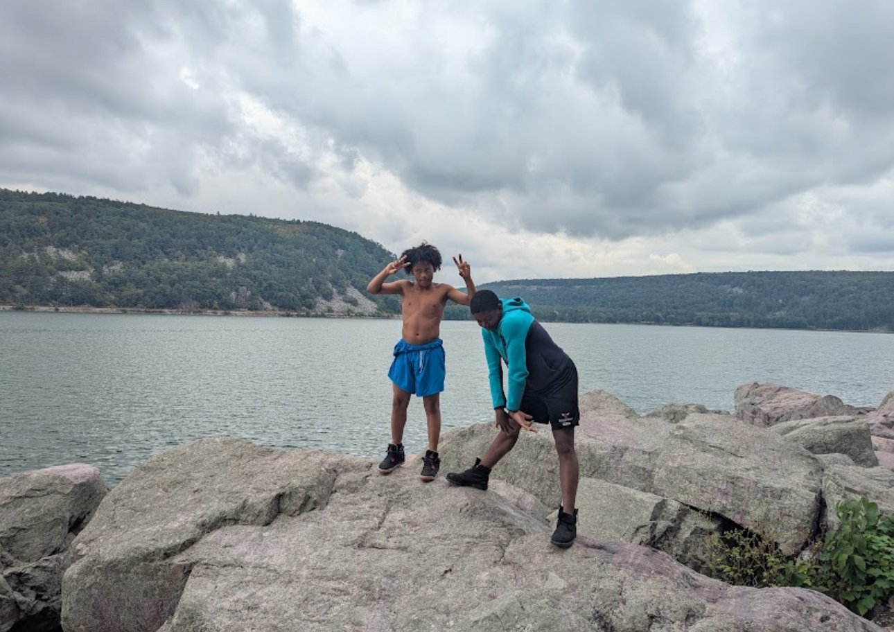 Two boys on large rocks near a lake with a cloudy sky and forested hills in the background.