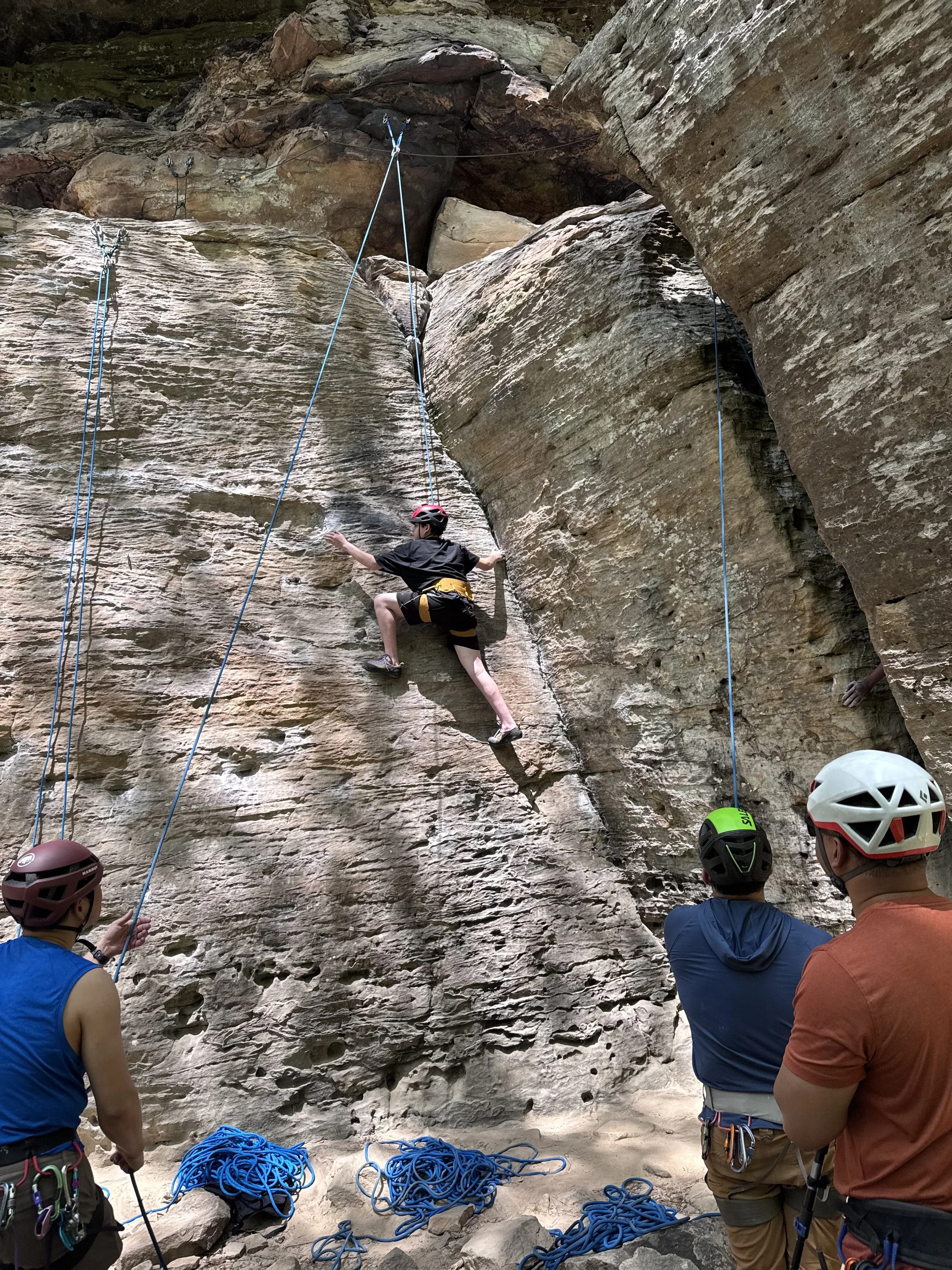 A group of rock climbers and onlookers gather at the base of a large outdoor rock face with climbing ropes, helmets, and gear, preparing for or taking a break from climbing.