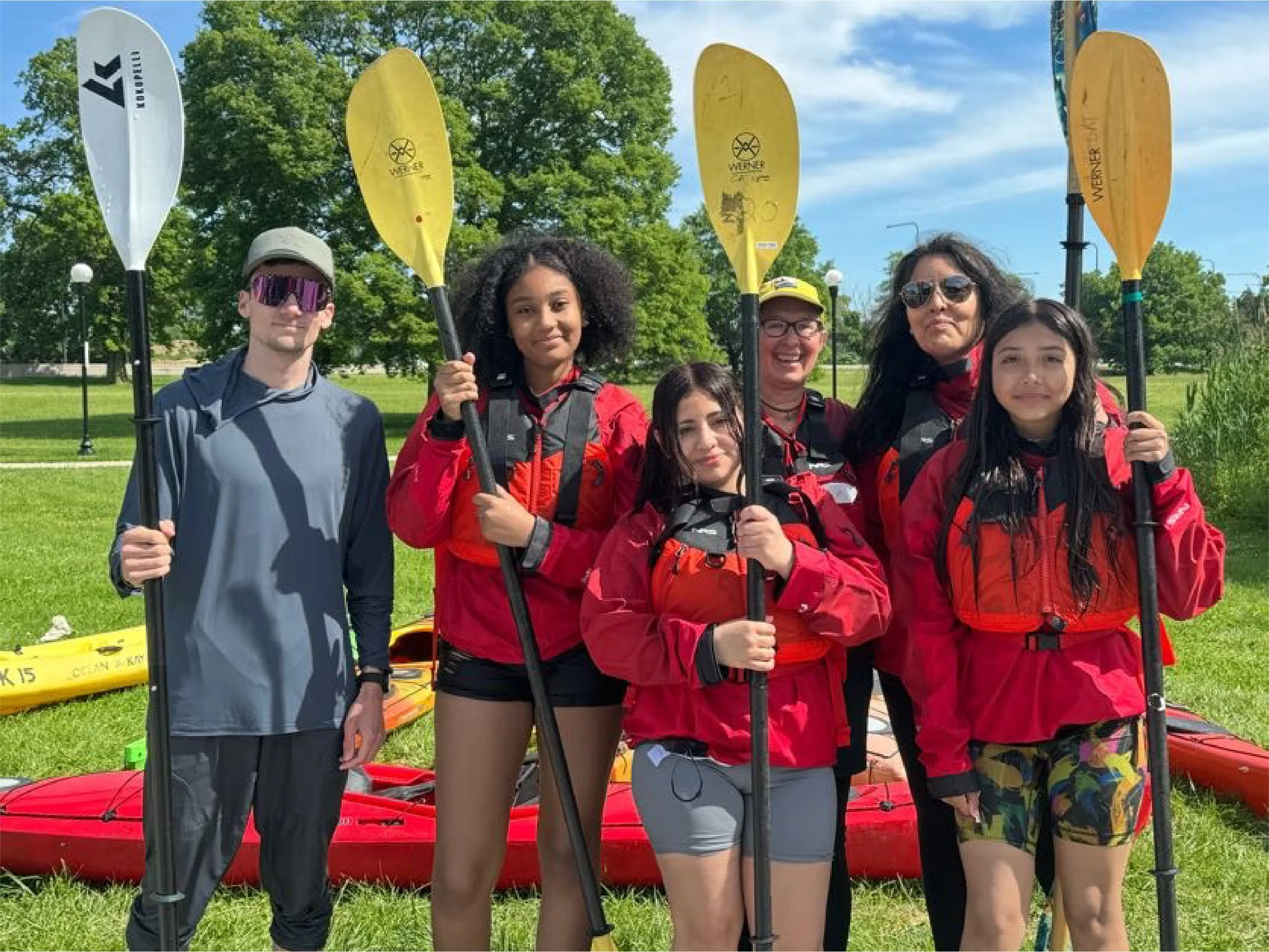 Group of six people holding paddles and standing in front of a red kayak, outdoors on a grassy area with trees and blue sky.