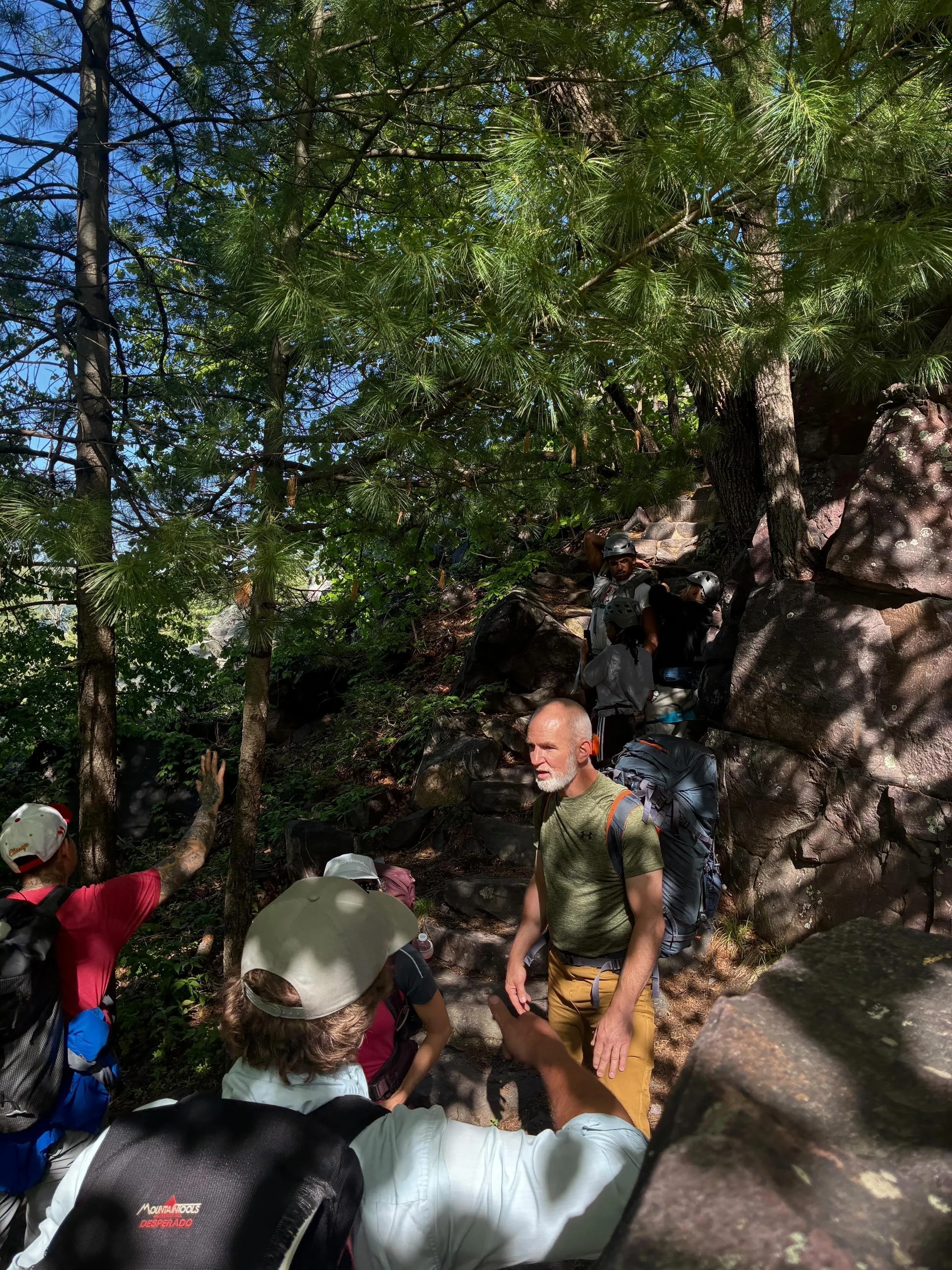 A group of people with backpacks and helmets hiking on a rocky trail in a dense forest with tall trees and green foliage, some people pointing or reaching out.
