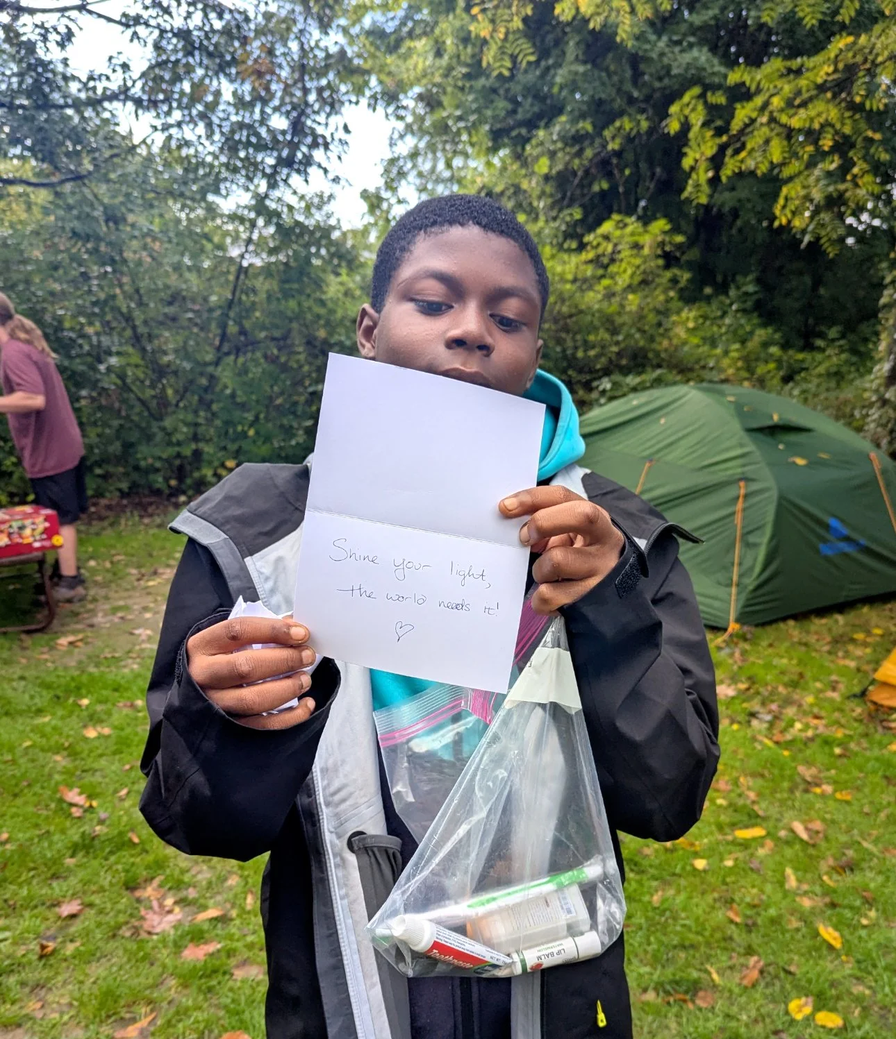A young boy in outdoor clothing holding a handwritten note that reads, 'Shine your light, the world needs it!' with a heart symbol. He is standing on grass with trees and a green tent in the background, and a person is partially visible on the left s