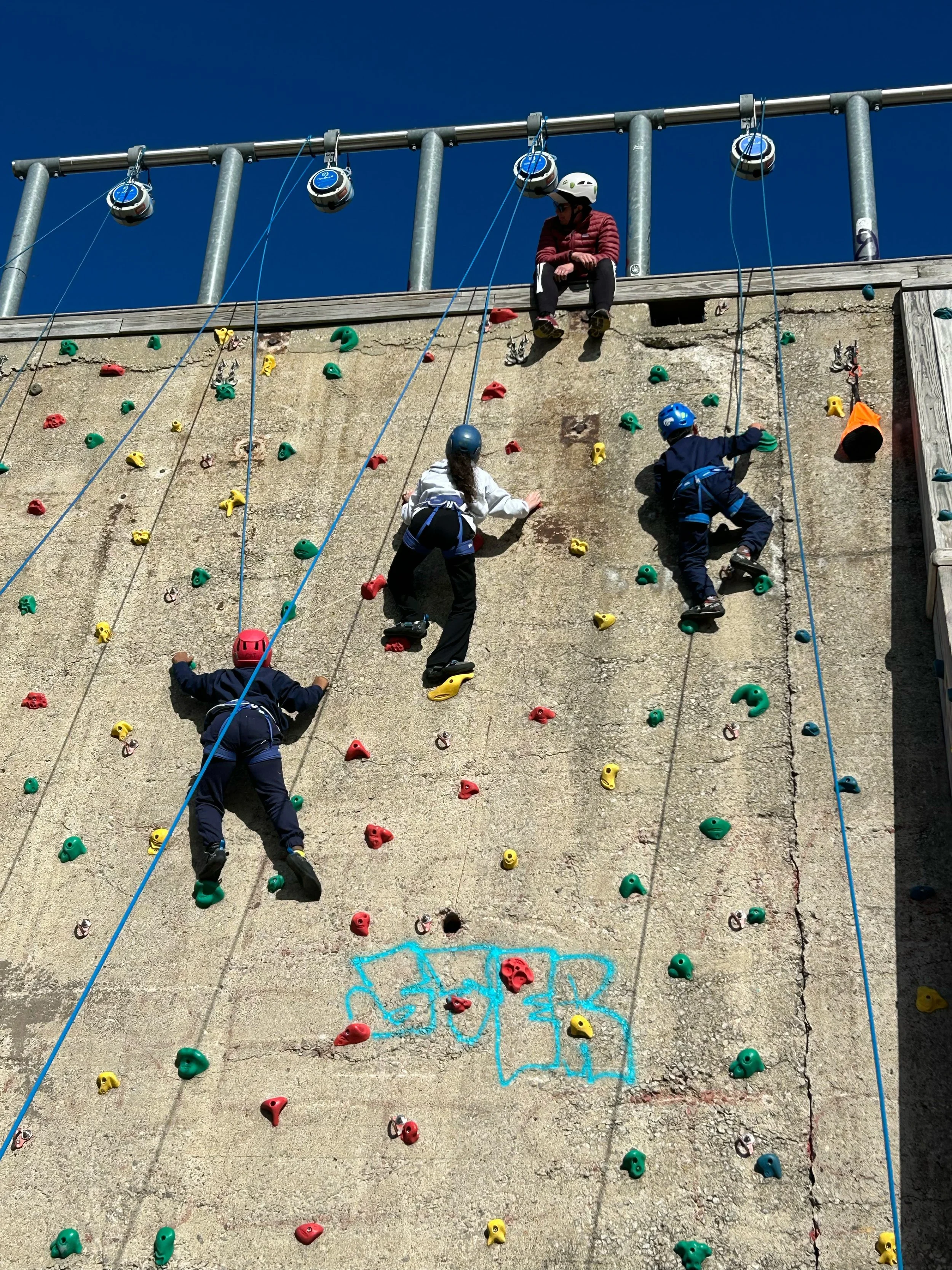 Kids climbing on an outdoor rock wall with colorful handholds, safety helmets, and harnesses, under a clear blue sky.