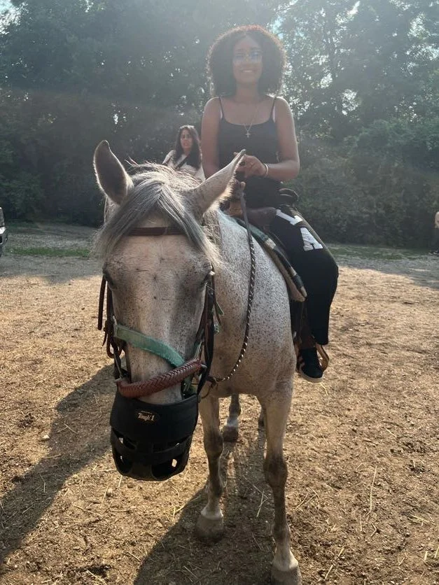 A woman with curly hair sitting on a white horse, wearing a black tank top, with another woman in the background standing in a wooded outdoor area.