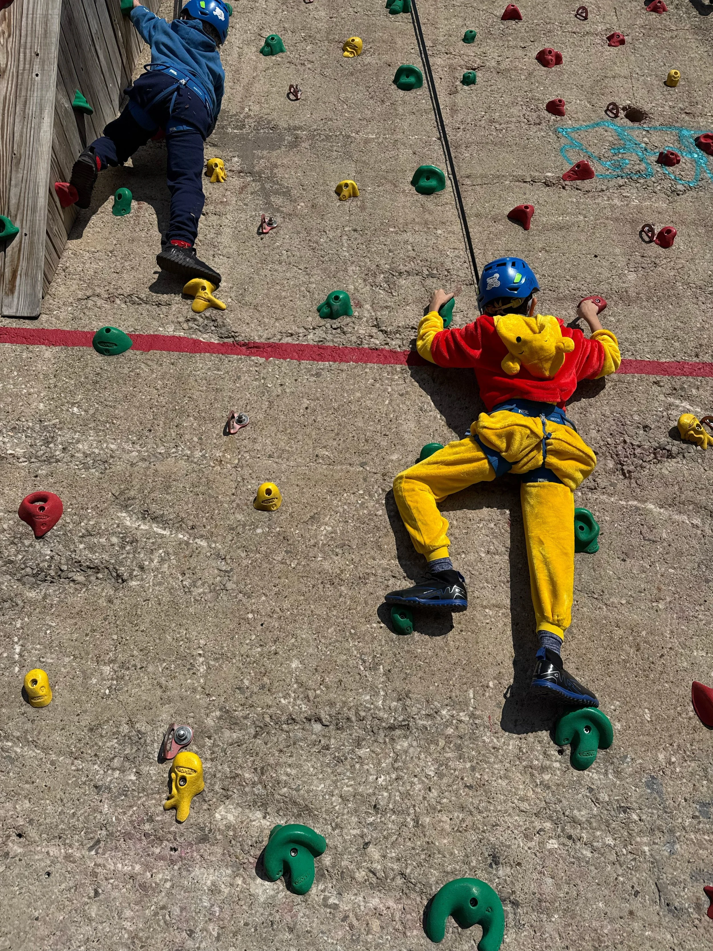 Two children wearing helmets and climbing harnesses practicing rock climbing on an outdoor artificial wall with colorful holds on a concrete surface.