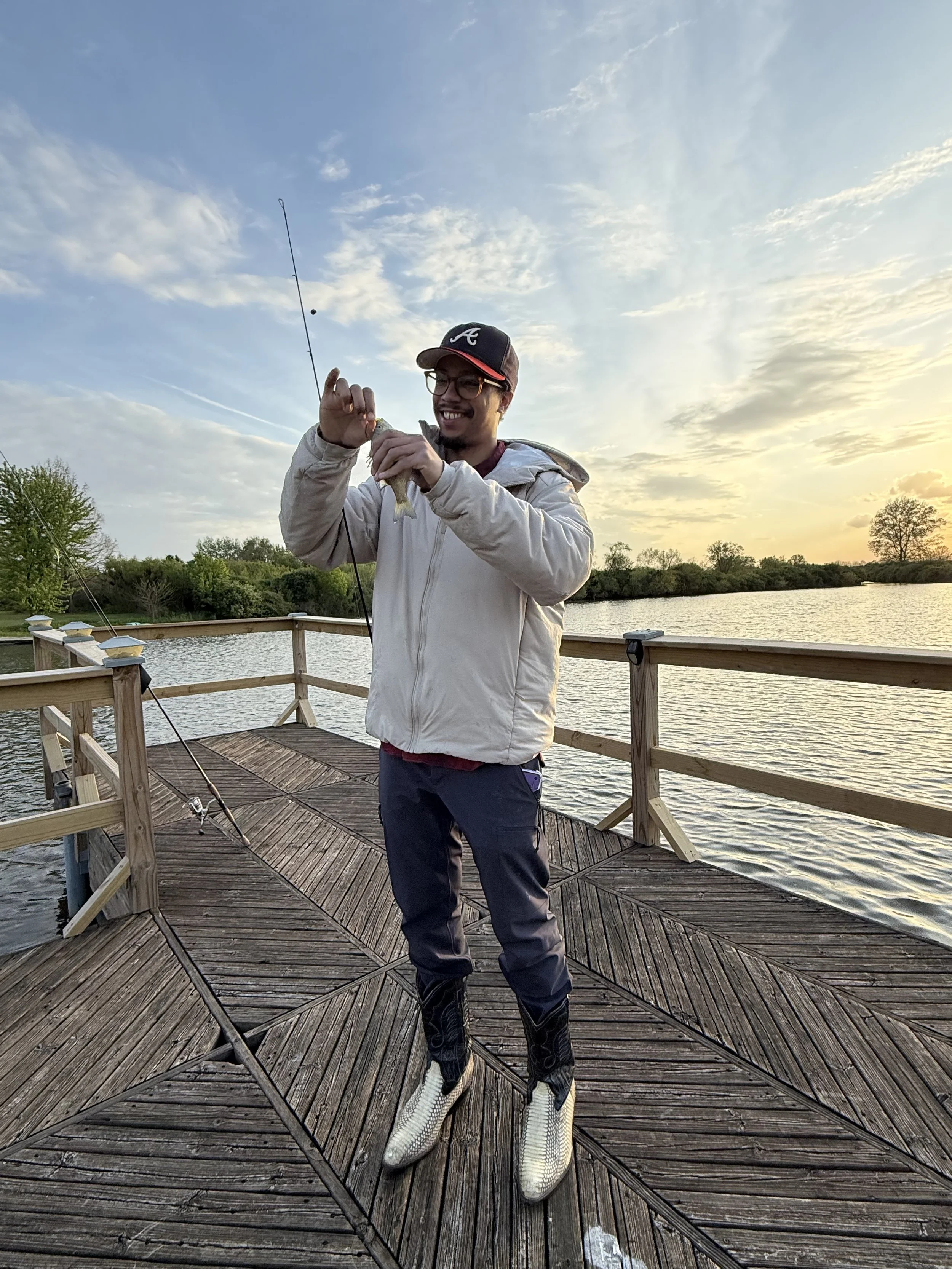 A man standing on a wooden dock holding a small fish in his hands, smiling, with a fishing pole nearby, water and trees in the background during sunset.