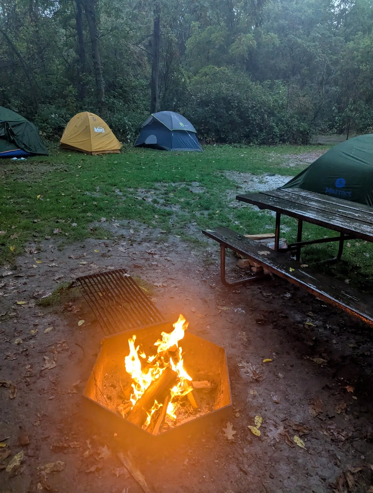 Camping scene at dusk with tents set up on grassy ground, a picnic table, and a fire pit with a fire in it. The background features trees and a wet, muddy ground.