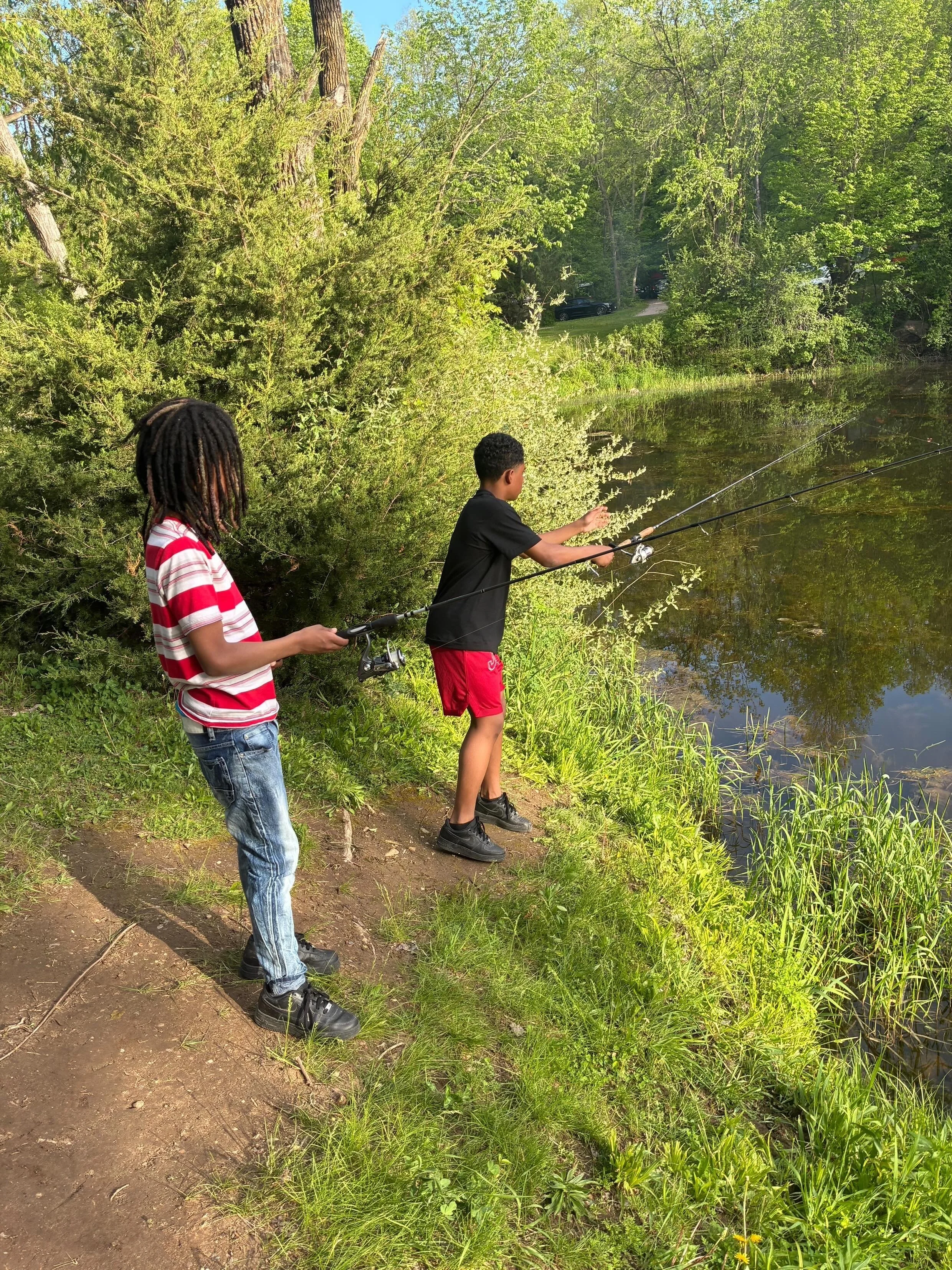Two children fishing by a pond in a lush, green outdoor setting on a sunny day.