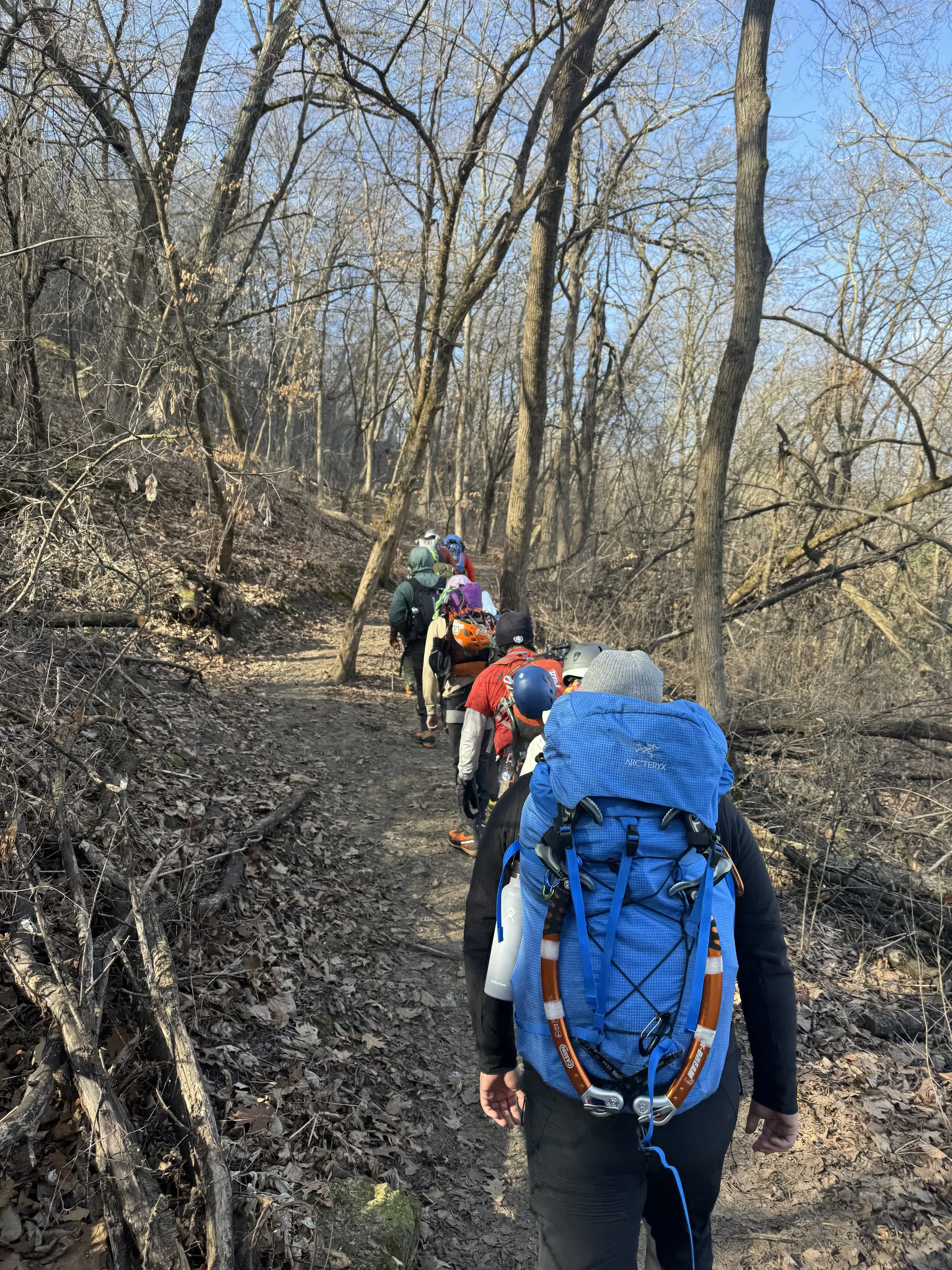 Group of hikers walking along a wooded trail on a clear day with leafless trees.
