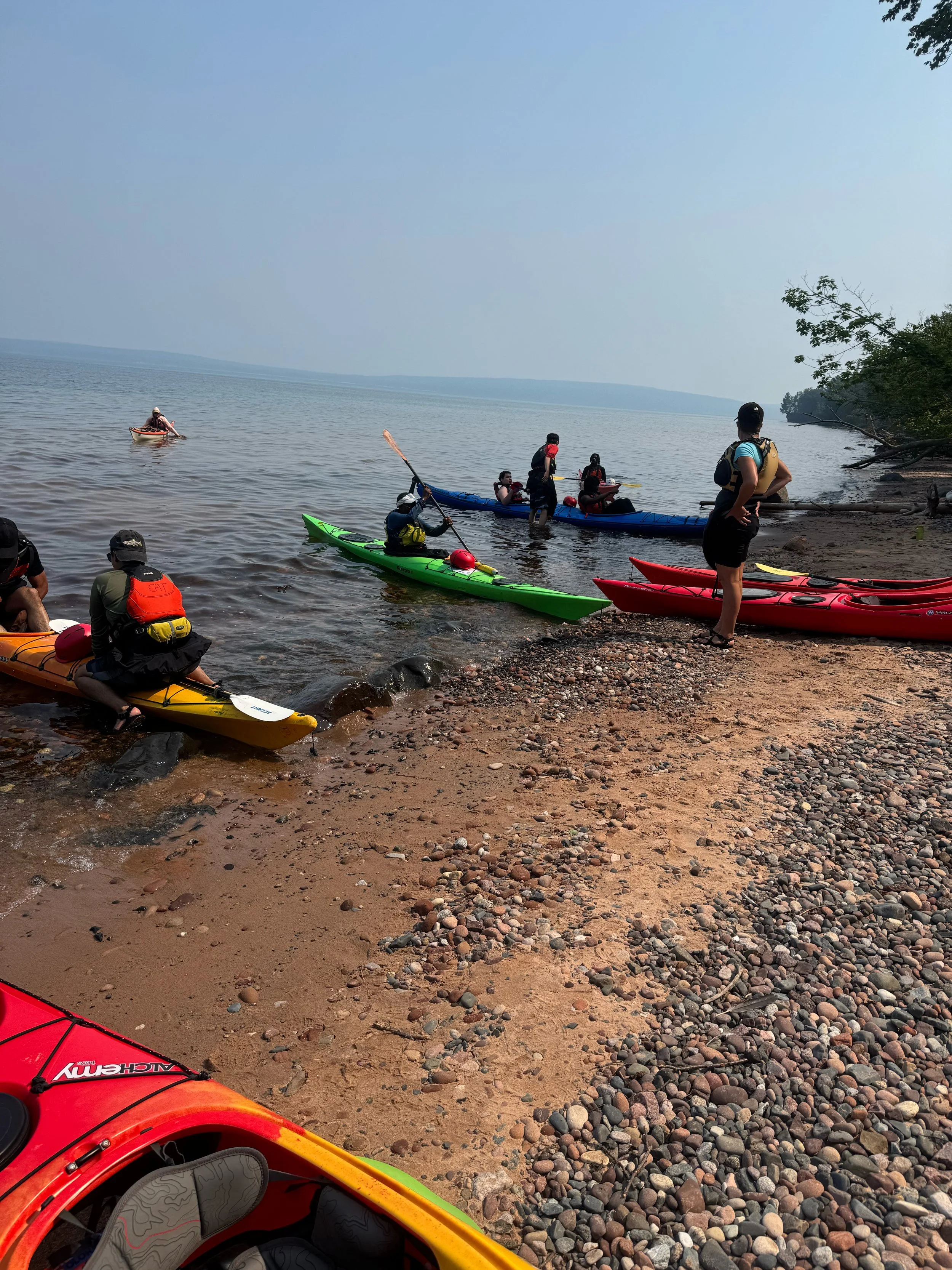 People preparing to kayak on a lake's shoreline with kayaks and paddles, some already in the water, surrounded by trees and rocks under a clear sky.