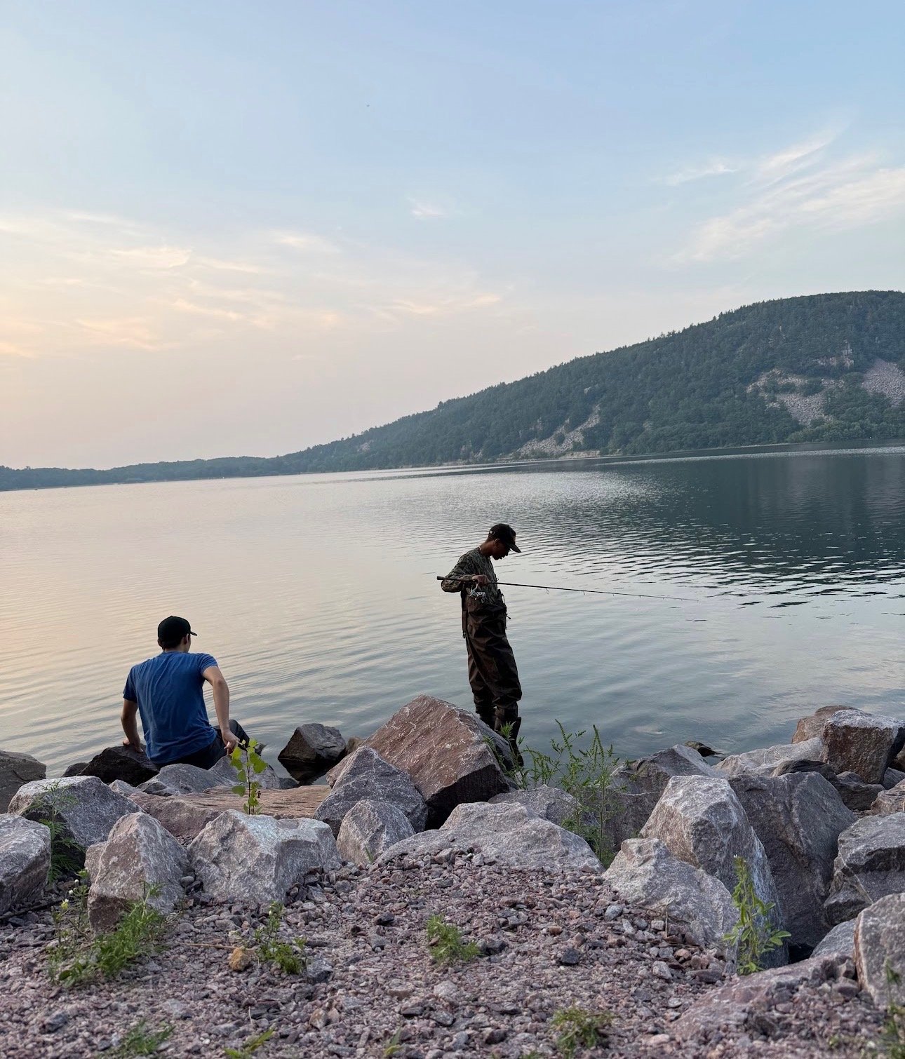 Two boys are by a lake with a wooded hillside in the background. One boy is sitting on rocks, wearing a blue shirt and a black cap. The other boy is standing on rocks, wearing a patterned shirt, dark pants, and a cap, holding a fishing rod.