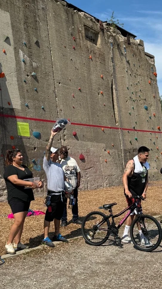 People standing near a large outdoor climbing wall with colorful holds. A man with a white towel around his neck is holding a helmet, with a bicycle nearby. Other individuals look on, some smiling, under a clear sky.
