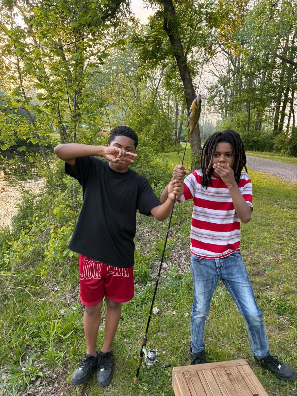 Two boys standing outdoors in a wooded area near a pond, holding a fishing rod with a fish hooked on it. The boy on the left is wearing a black T-shirt and red shorts, making a peace sign with his right hand. The boy on the right is wearing a red and