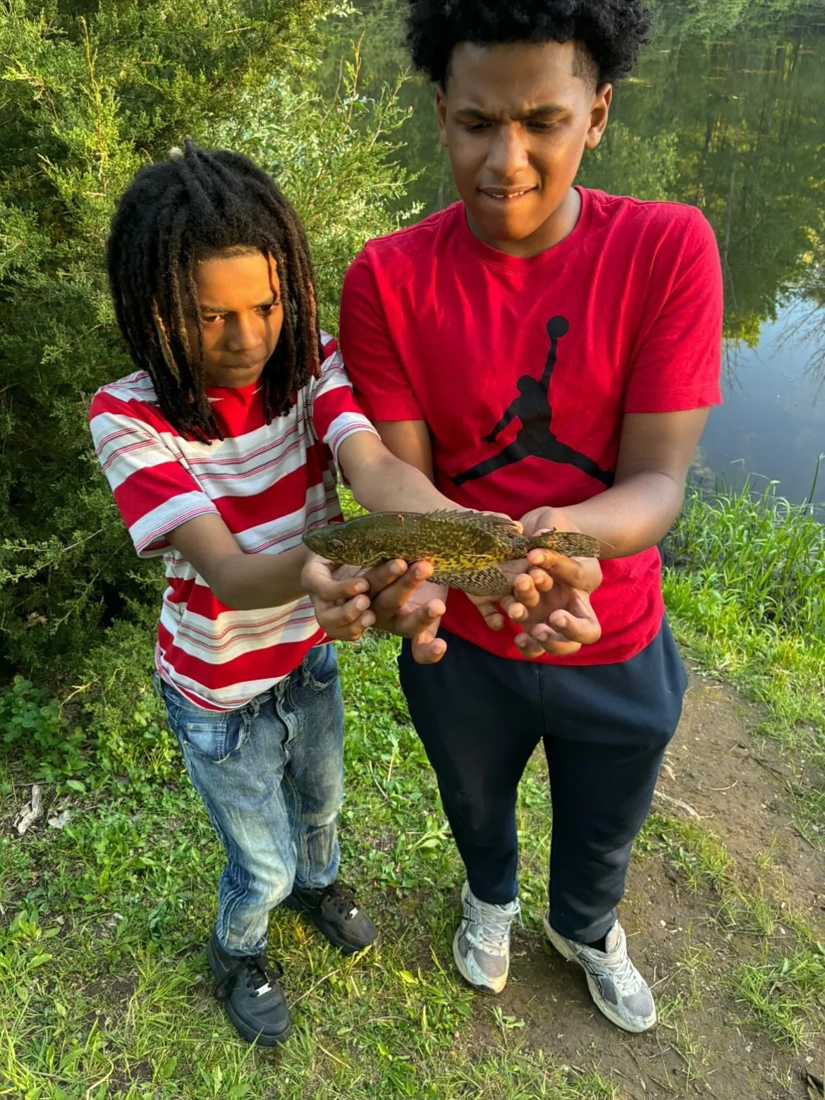 Two boys by a pond holding a fish, with trees and water in the background.