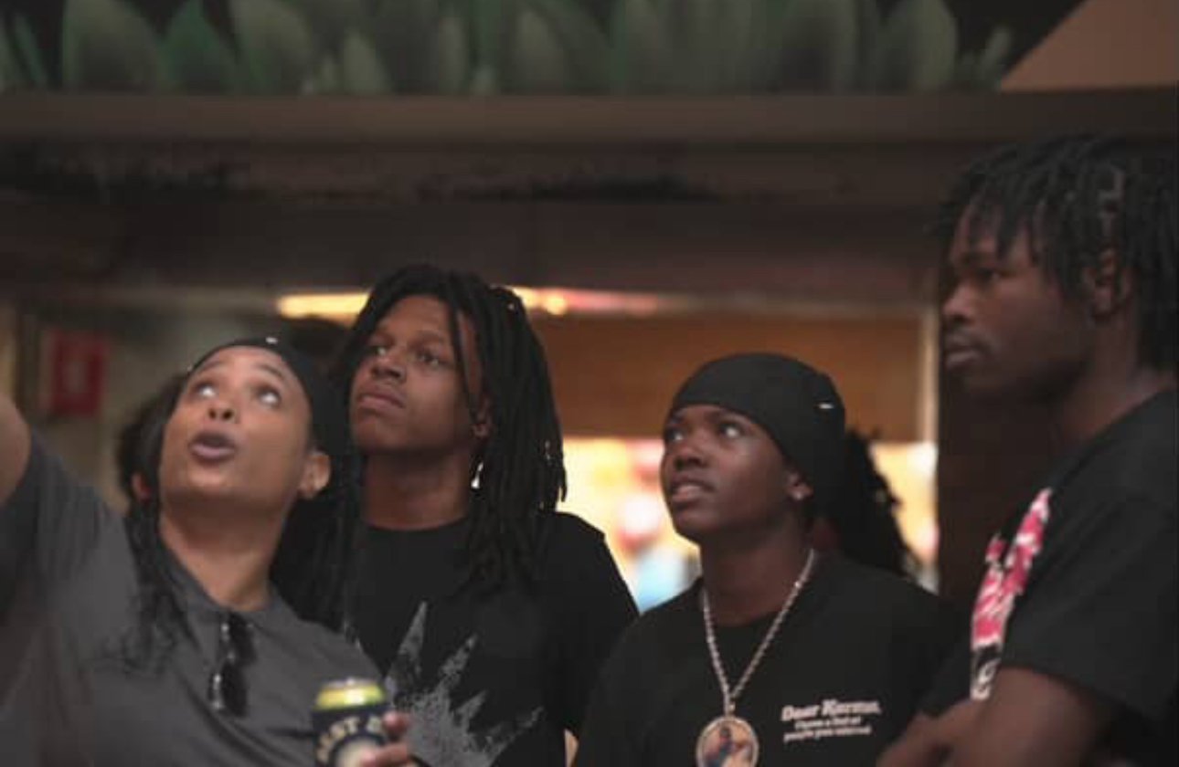 Four young women standing together indoors, one on the far left holding a drink can and pointing upward, with a woman in a gray shirt, another with dreadlocks, another with a black bandana and a necklace, and a tall man with dreadlocks on the far right, all looking intently in the same direction.