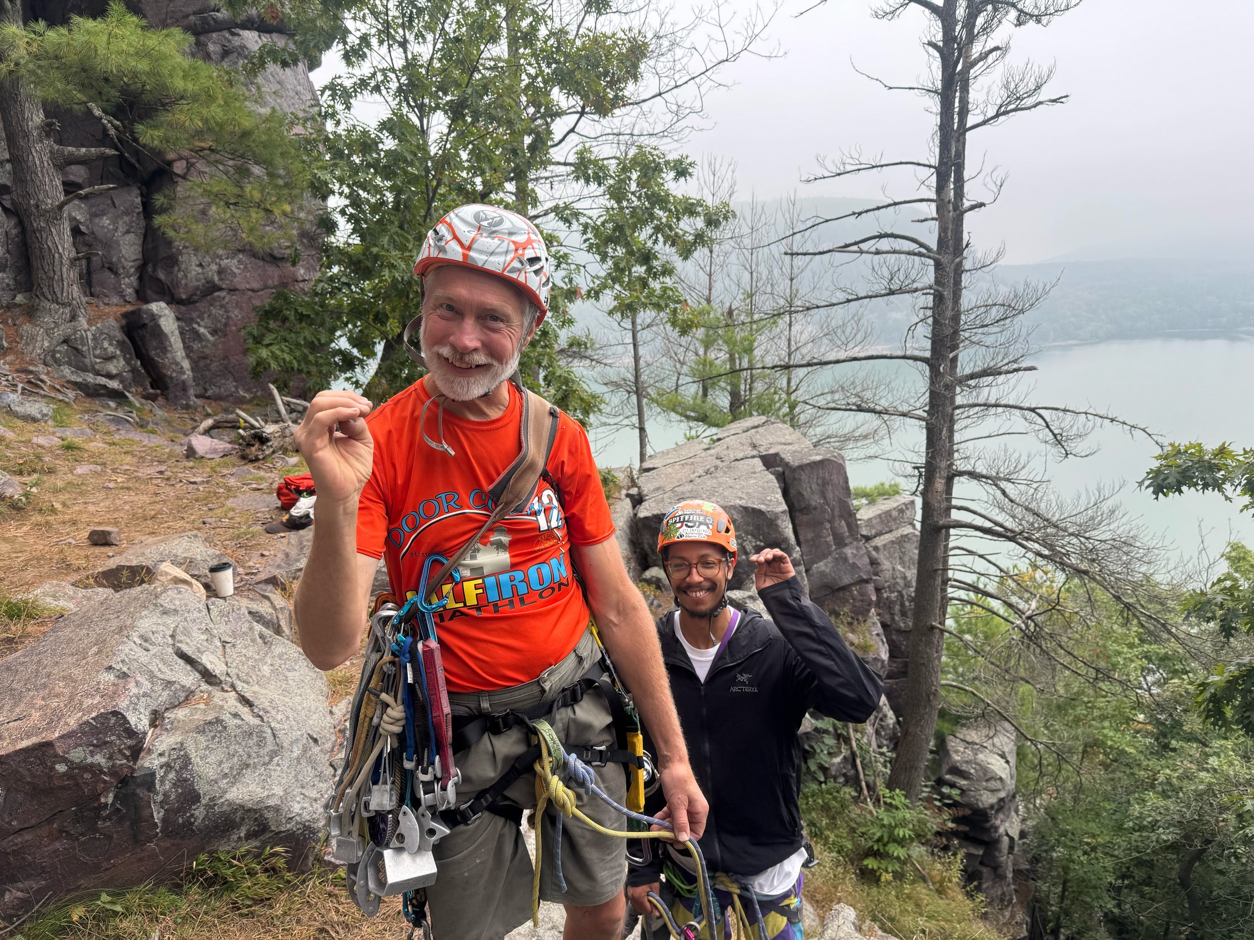 Two smiling men hiking on a rocky trail near a lake, surrounded by trees and mountains, with overcast skies.