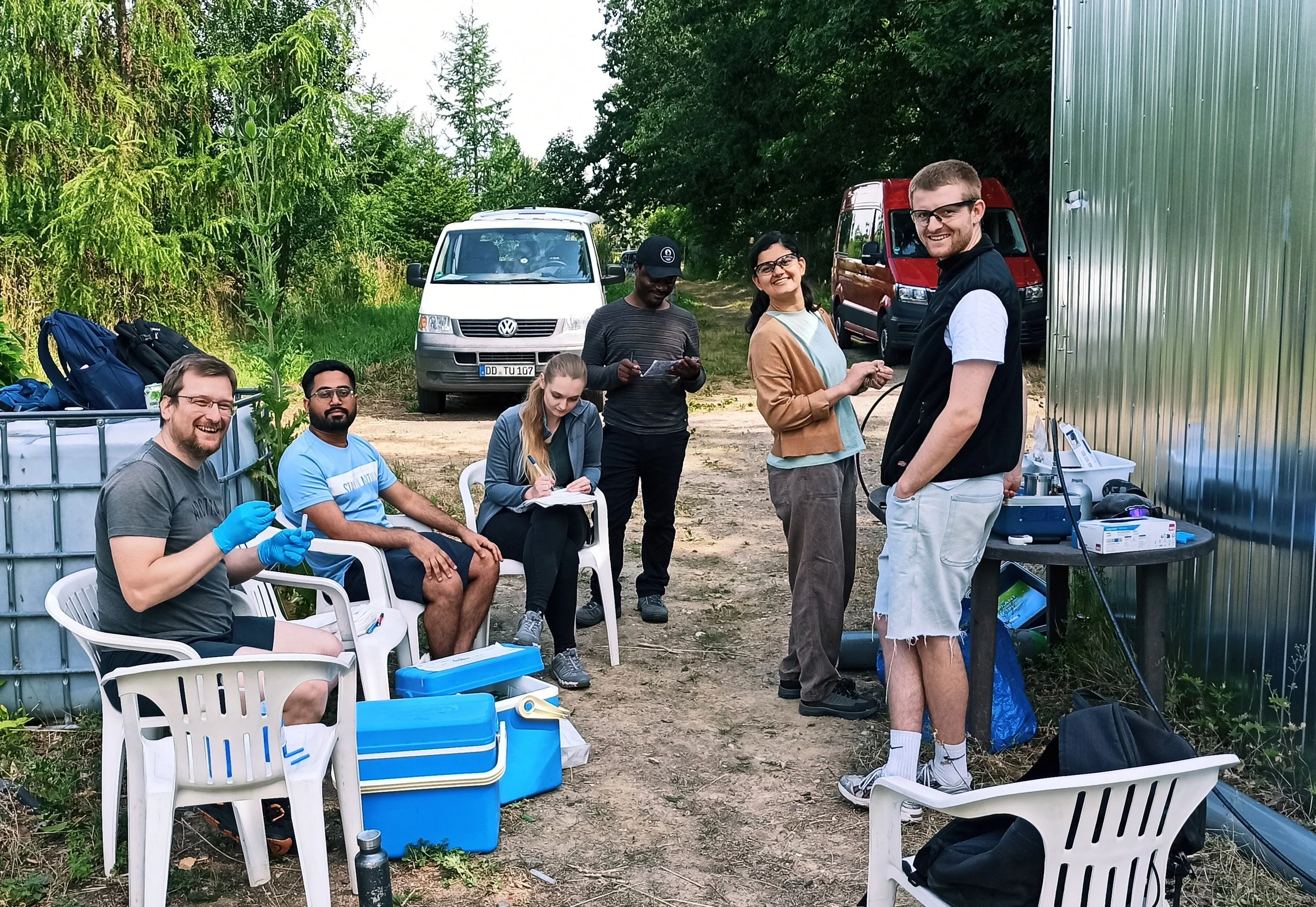 Group of young researchers outdoors working with scientific equipment, some seated on chairs, others standing near a table, with vehicles and greenery in the background.