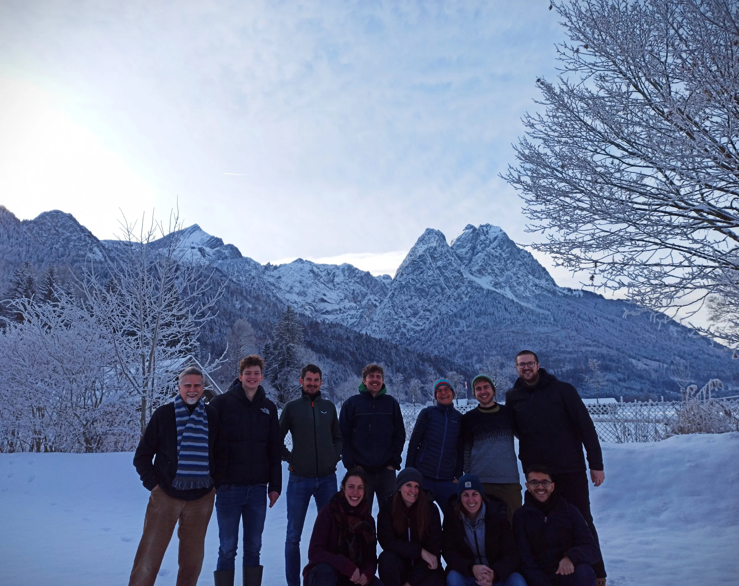 Research team standing and sitting in snow with mountains and a cloudy sky in the background during winter.