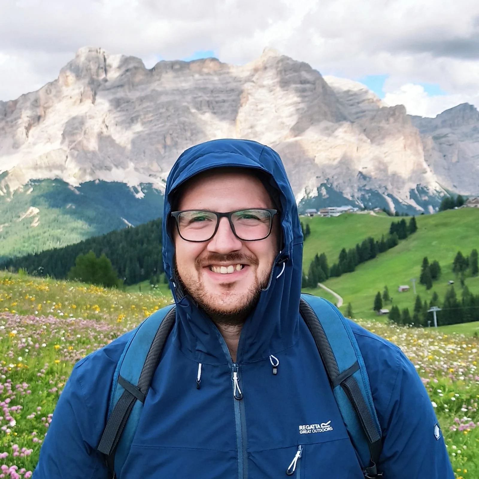 Scientist in blue outdoor jacket and glasses, standing in a lush green meadow with colorful wildflowers, with mountains and cloudy sky in the background.