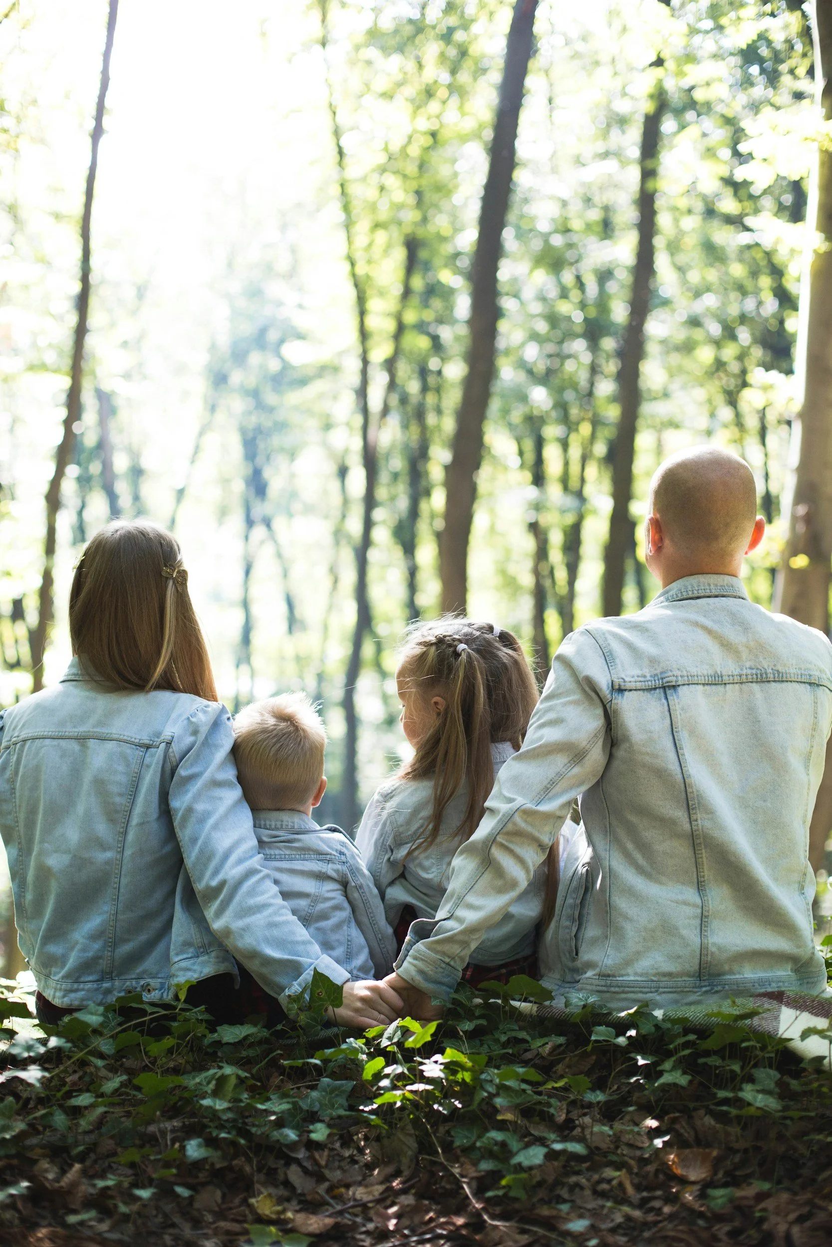 A family of five sitting on the ground in a sunny forest, with their backs to the camera. The family includes two adults and three children, all wearing light-colored denim jackets.