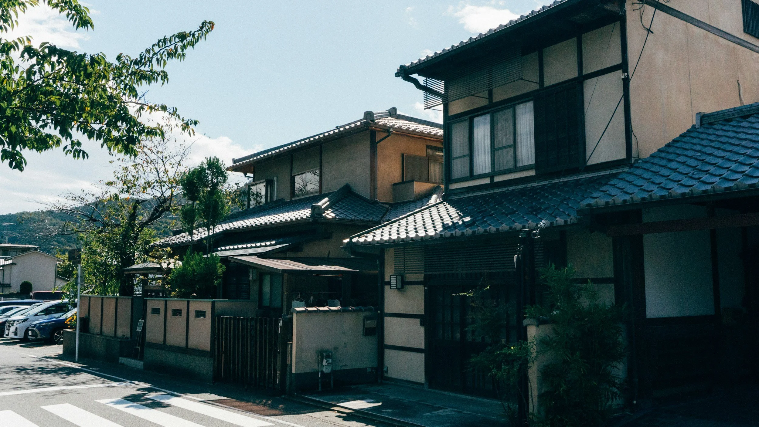 A street view of traditional Japanese-style houses with tiled roofs, a wooden fence, and a parking area with cars, under a partly cloudy sky with trees in the foreground.