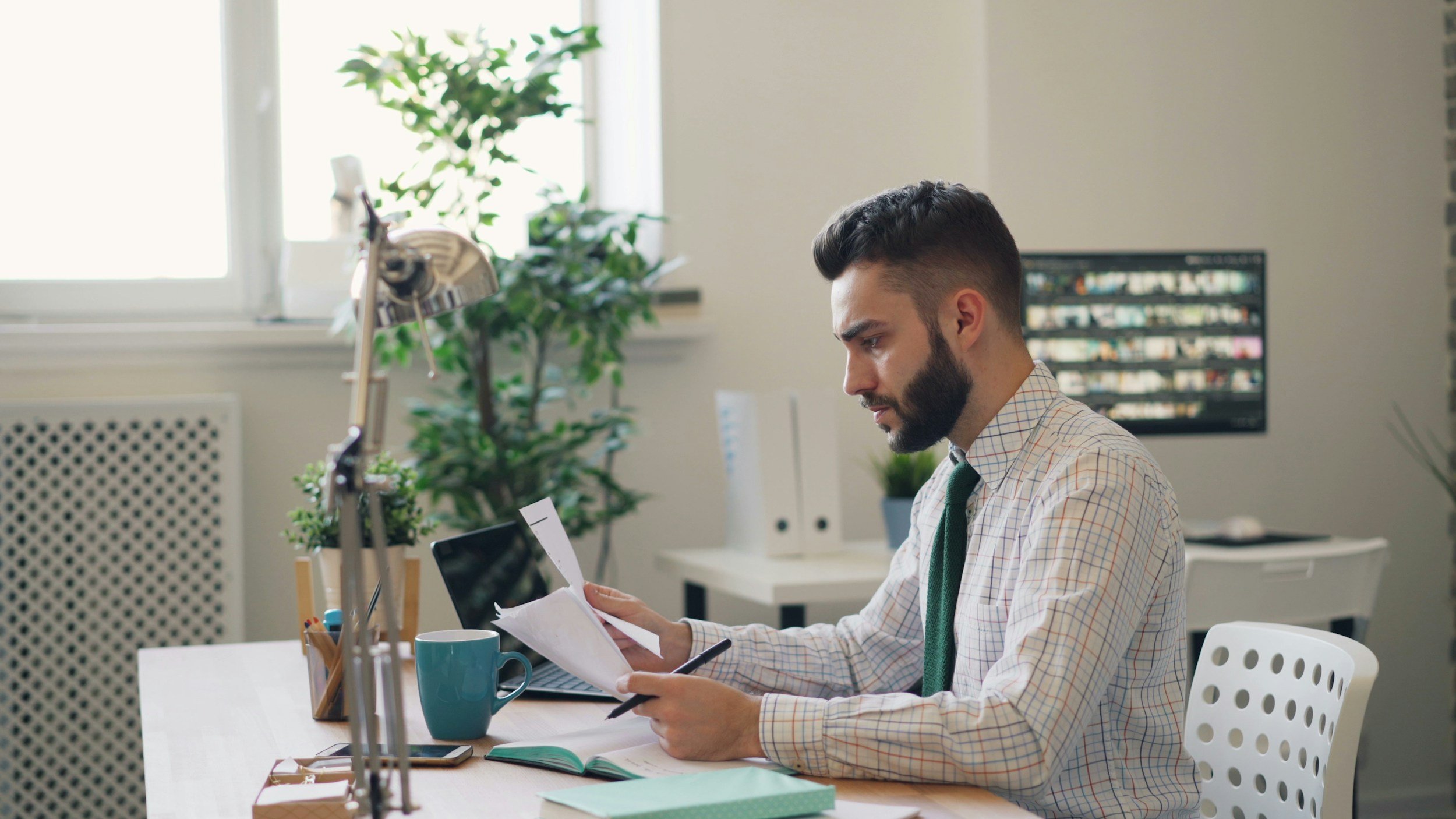 Man sitting at desk reading papers with a coffee mug, laptop, and office supplies, in an office environment with plants and a computer monitor in the background.