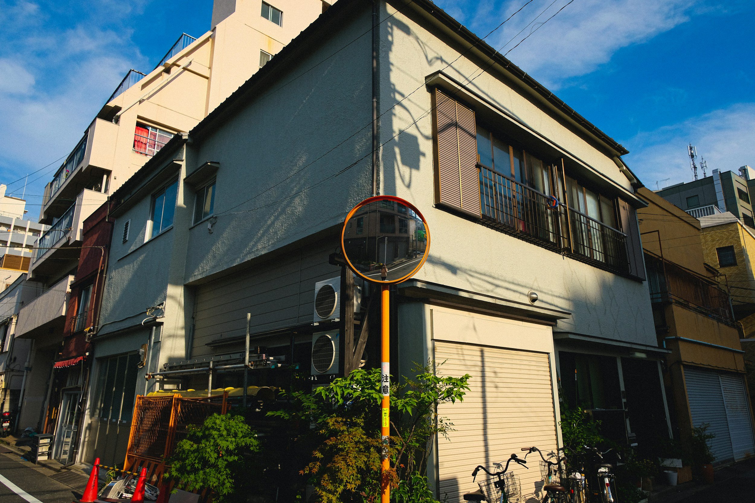 A corner building on a city street, with various residential and commercial units, a convex street mirror reflecting the street, bicycles parked outside, and shadows cast by nearby trees and power lines.