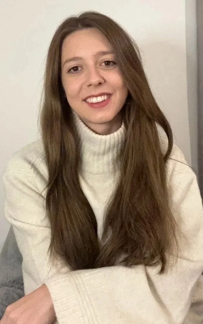 Young woman with long brown hair wearing a cream turtleneck sweater, smiling at the camera.