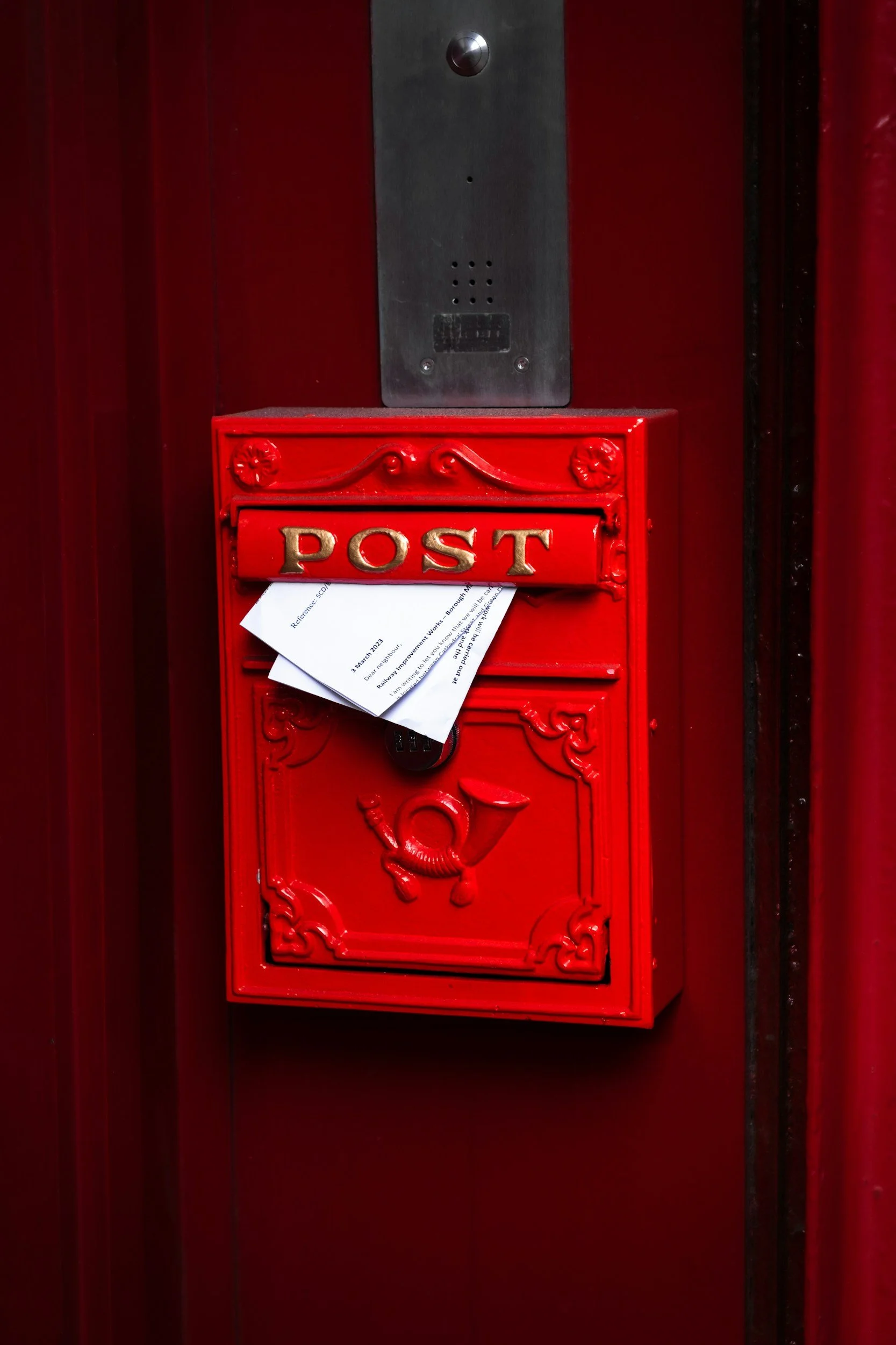 A red mailbox with a slot labeled 'POST' on the front, mounted on a red wall, with some papers and a lock visible inside the slot.
