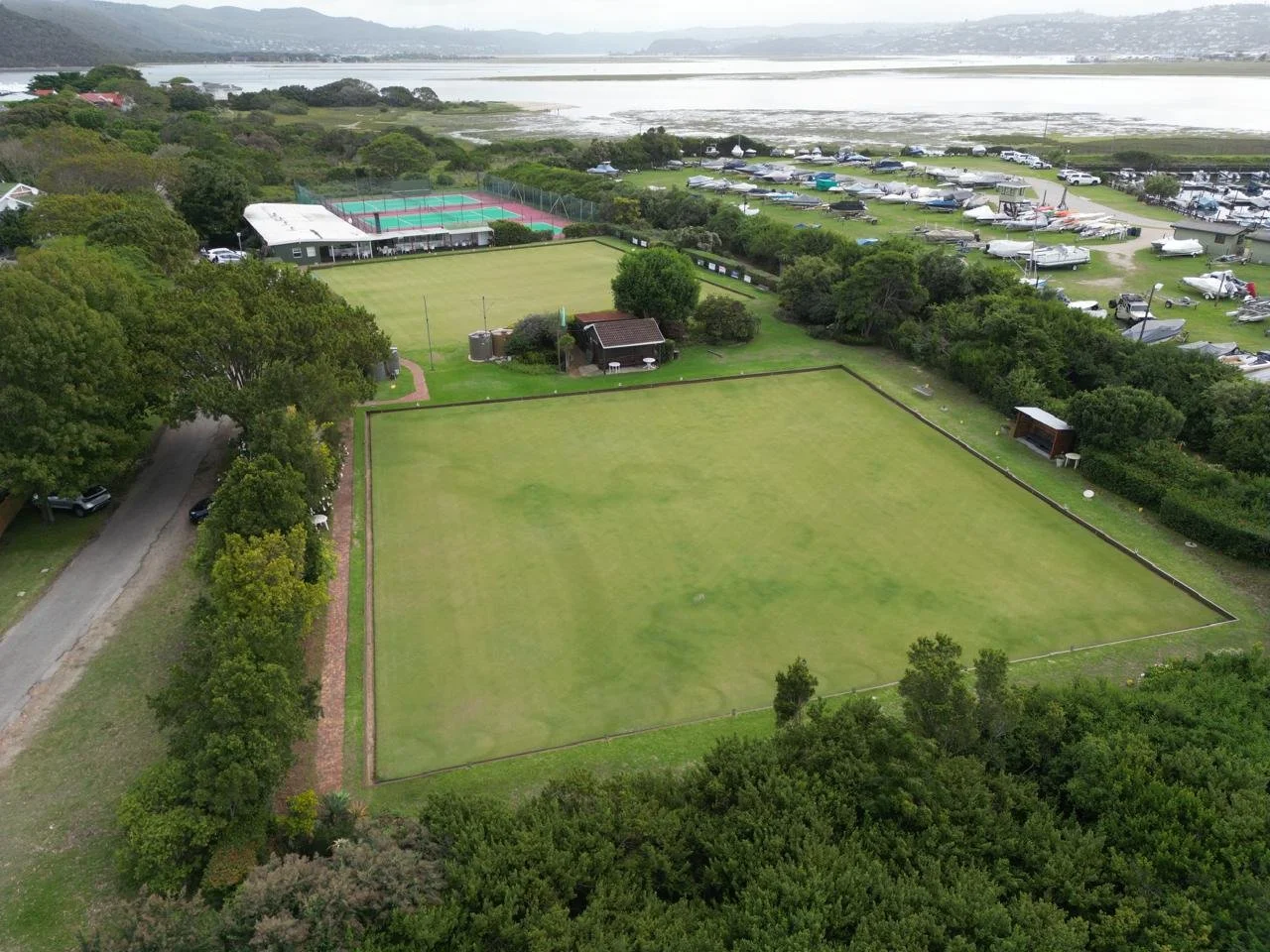 Aerial view of two green lawn sports fields, tree-lined surrounding area, parking lot with boats and buses, marina by the water, and distant mountains.
