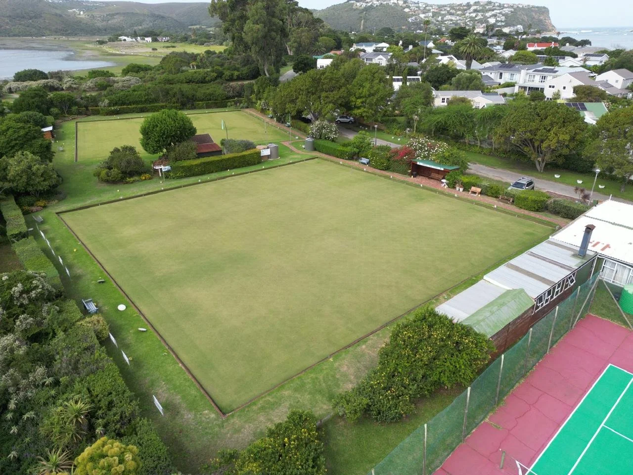 An aerial view of a large grass sports field, surrounded by trees, buildings, and houses, with a tennis court and water in the background.
