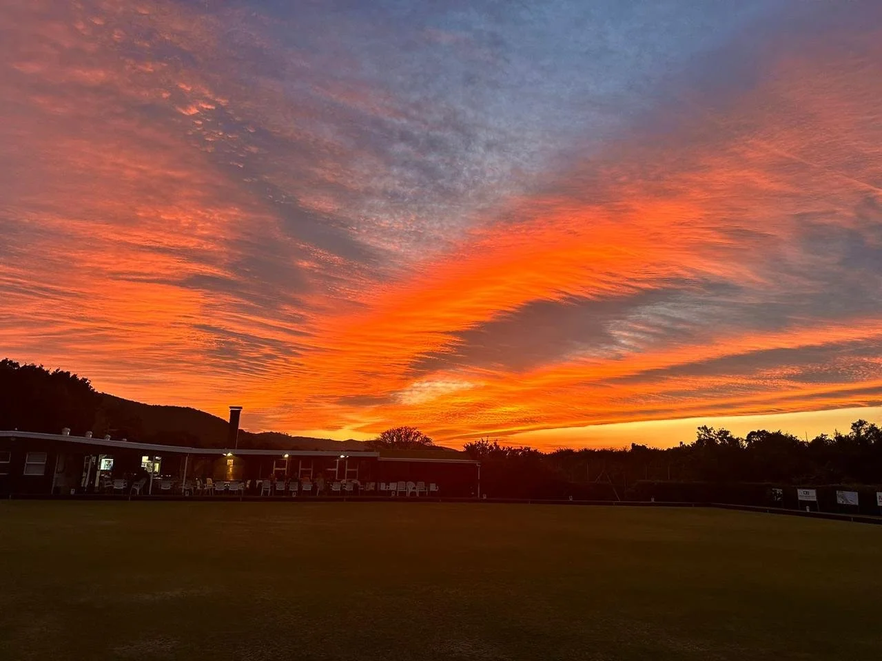 Sunset sky with vibrant orange and pink clouds over a grassy field, with a building and trees in silhouette.