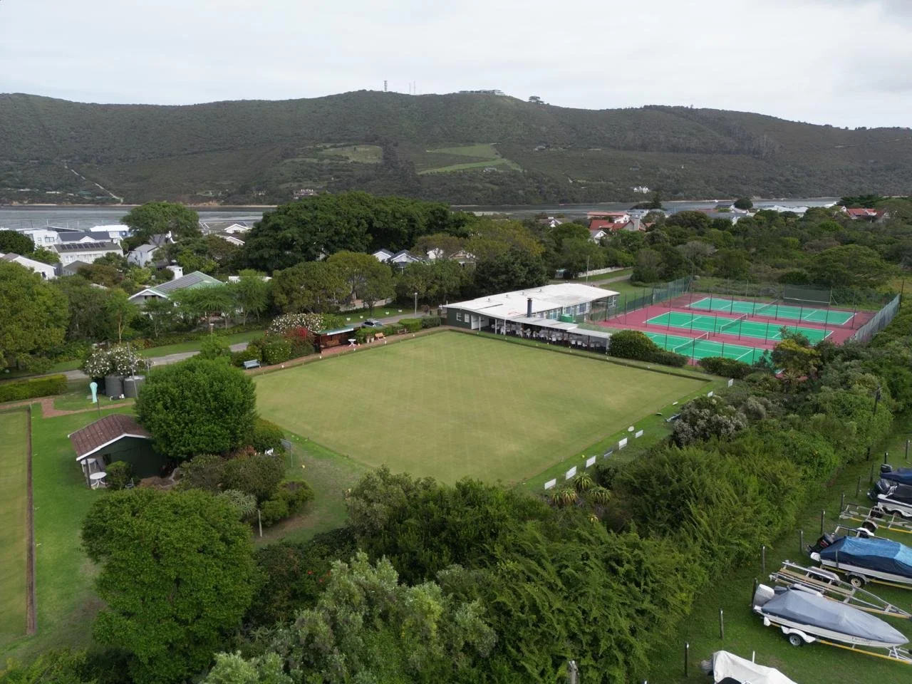 Aerial view of a recreational area with a large grass field, tennis courts, a building, parking lot, trees, and surrounding houses and hills in the background.